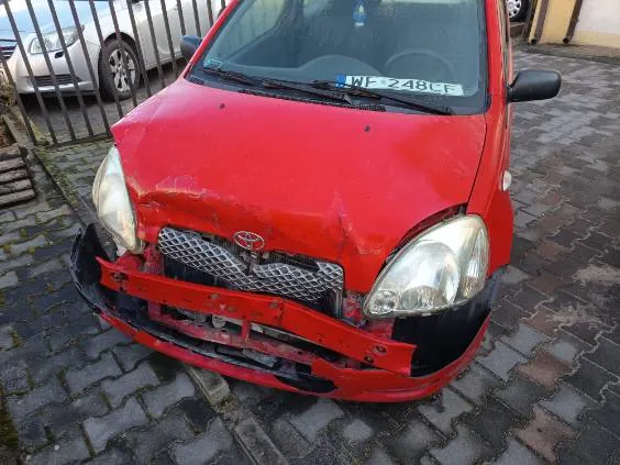 Czerwony samochód marki Toyota z widocznymi uszkodzeniami przedniej części: zgnieciona maska, uszkodzony zderzak i atrapa chłodnicy. Na przedniej szybie widoczny numer rejestracyjny WP 248CT. W tle metalowe ogrodzenie, częściowo widoczny srebrny samochód oraz brukowana powierzchnia. Samochód stoi na brukowanej nawierzchni.