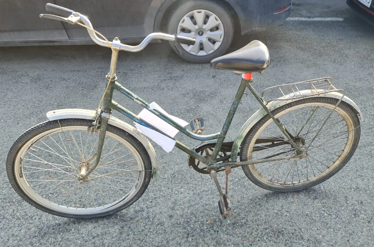 Zielony rower z białymi błotnikami, czarnym siedziskiem i metalowym bagażnikiem na tylnej ośce. Na ramie roweru przyczepiony jest biały arkusz papieru. W tle widoczny fragment ciemnego samochodu z kołem o srebrnej felgach. Rower stoi na asfaltowej powierzchni.