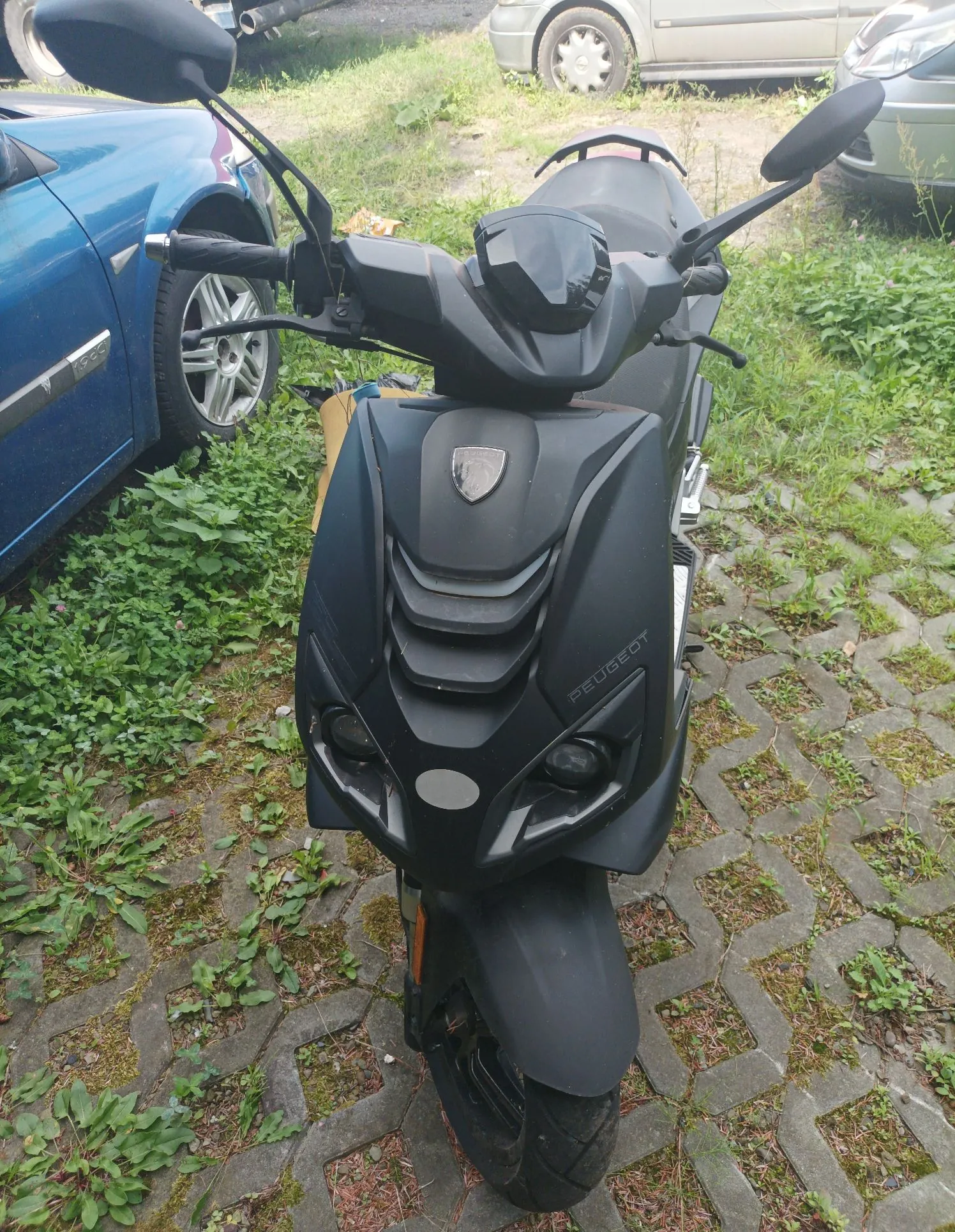 Widok z przodu na czarny skuter marki Peugeot, zaparkowany na kostce brukowej z trawą w szczelinach. Skuter ma charakterystyczny przedni pas z logo marki oraz napisem PEUGEOT po prawej stronie. W tle widoczny jest fragment niebieskiego samochodu po lewej stronie oraz jasne samochody w oddali.