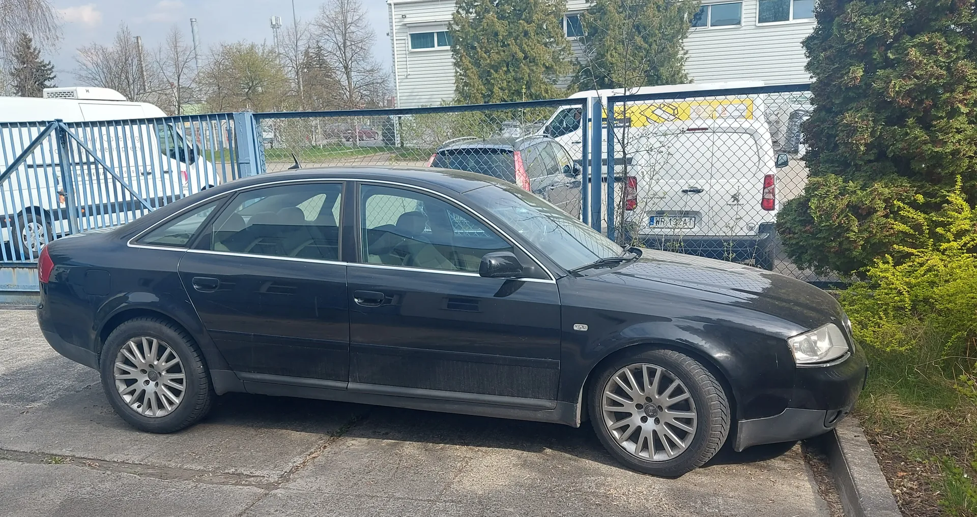 Czarny samochód osobowy typu sedan zaparkowany na betonowej powierzchni. W tle metalowe ogrodzenie siatkowe, biała furgonetka z tablicą rejestracyjną WR 132AT oraz żółtym napisem na dachu, niebieska brama, drzewa i budynek z białymi panelami. Po prawej stronie zielone krzewy i trawa. Samochód ma srebrne felgi i ciemne szyby.