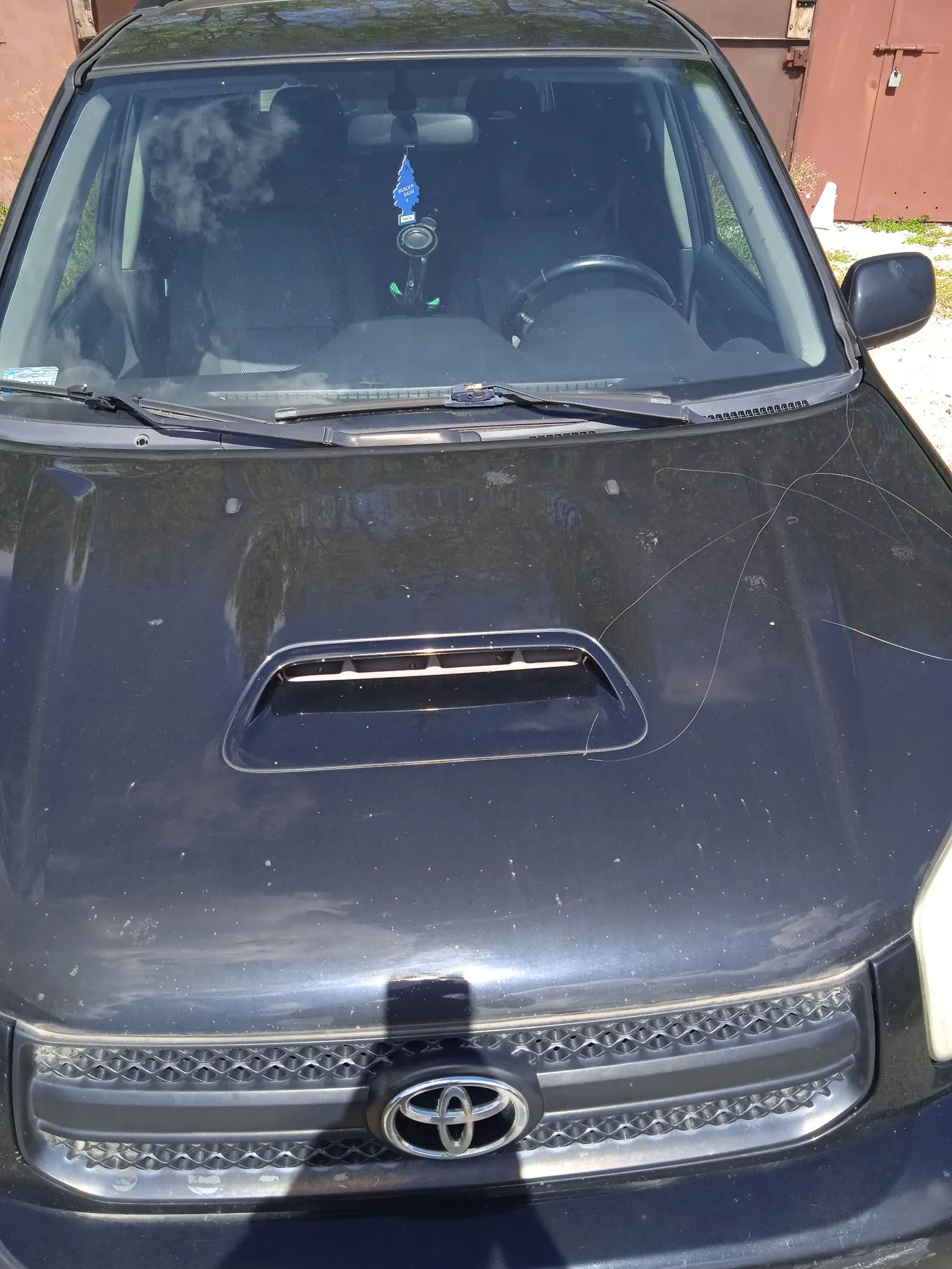Widok z przodu na czarny samochód marki Toyota. Widoczna jest maska z wlotem powietrza, grill z logo Toyoty na środku oraz przednia szyba, przy której lusterku wisi niebieski zapach samochodowy.