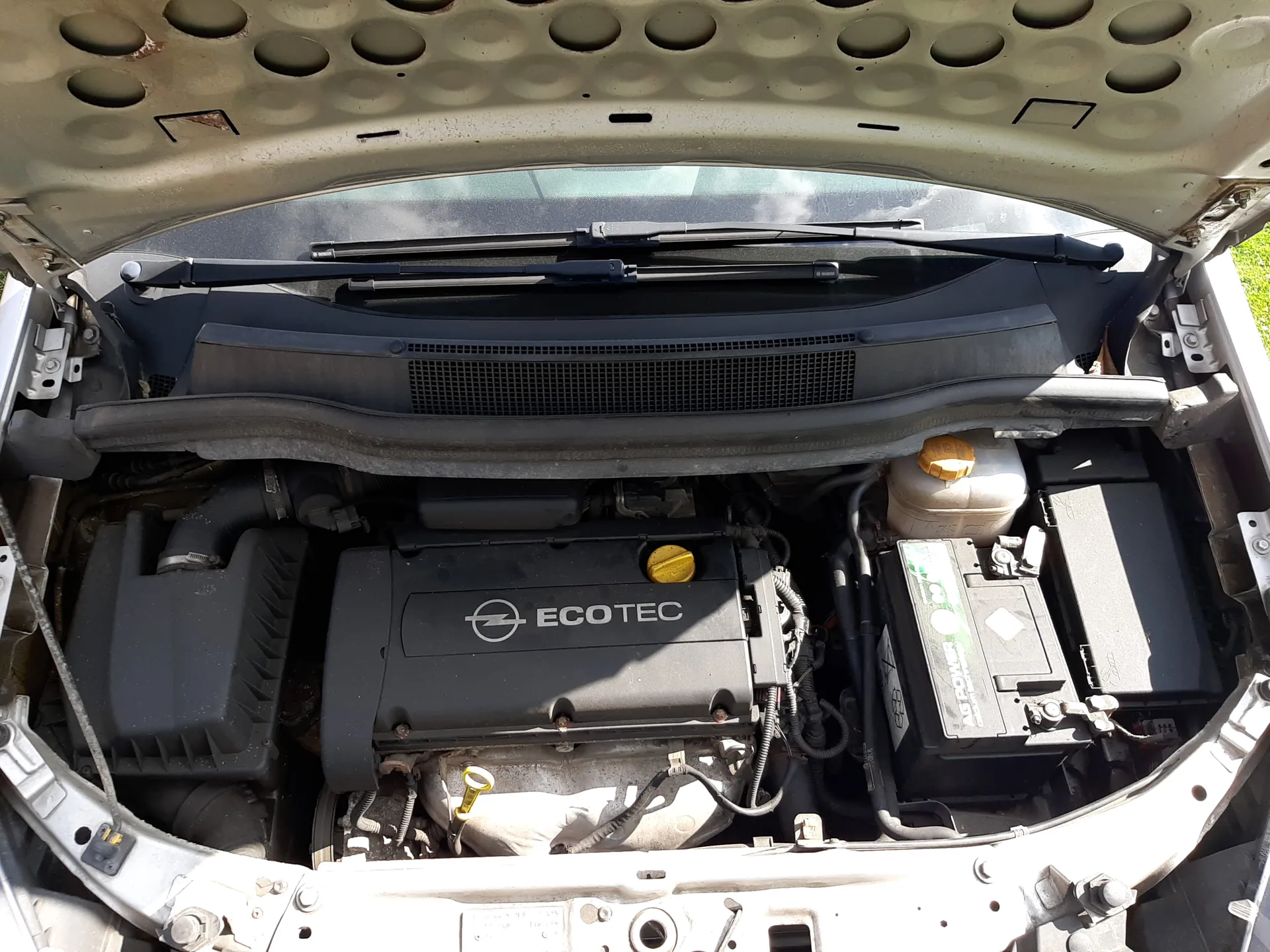 Widok na otwarty maskę samochodu z silnikiem. Centralnie umieszczona czarna pokrywa silnika z napisem ECOTEC i logo. Po prawej stronie bateria z zielonymi i białymi elementami oraz zbiornik chłodniczy z pomarańczową nakrętką. W tle przednią szybę z wycieraczkami i siatkę wentylacyjną. Maskę charakteryzuje wzór okrągłych wgłębień. Wszystkie elementy są wyraźnie widoczne i prawidłowo ustawione.
