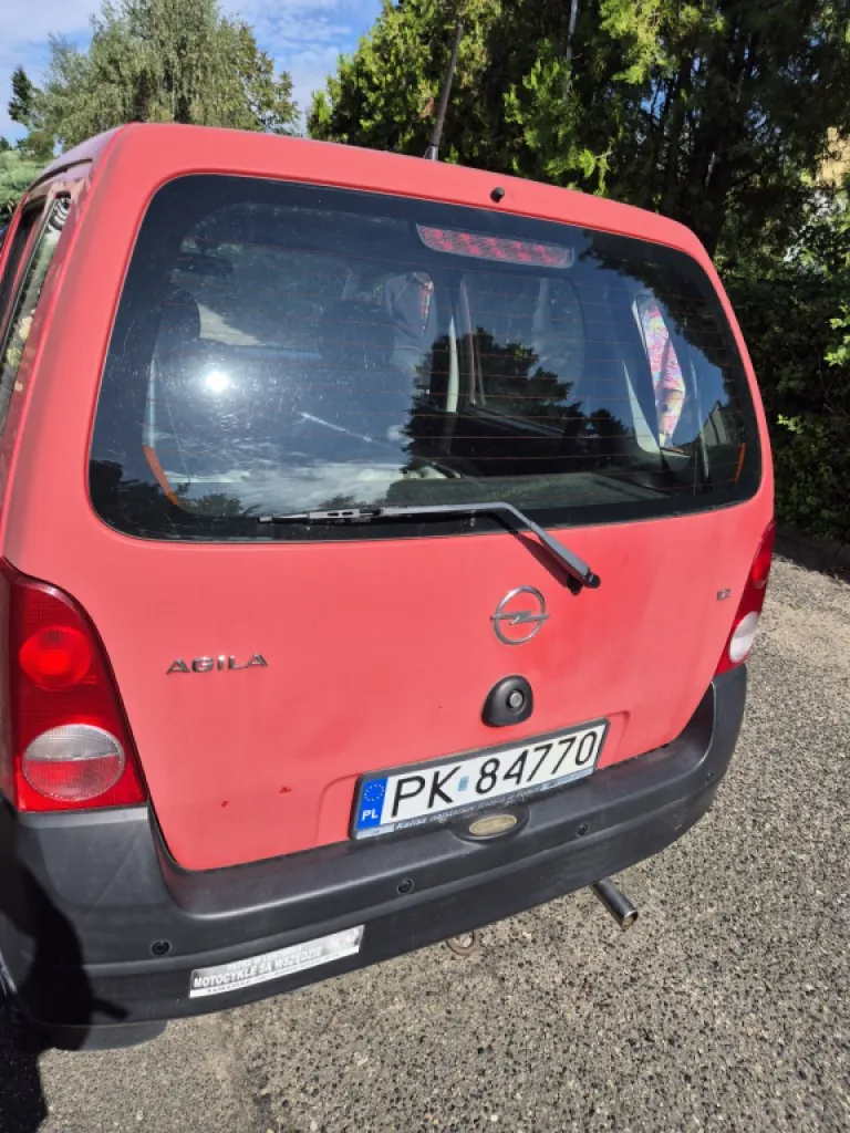 Tylny widok czerwonego samochodu marki Opel Agila. Na klapie bagażnika widoczne jest logo Opela oraz napis 'AGILA'. Samochód posiada polską tablicę rejestracyjną o numerze PK 84770. Pojazd stoi na żwirowym podłożu, w tle widoczne są zielone drzewa i krzewy.