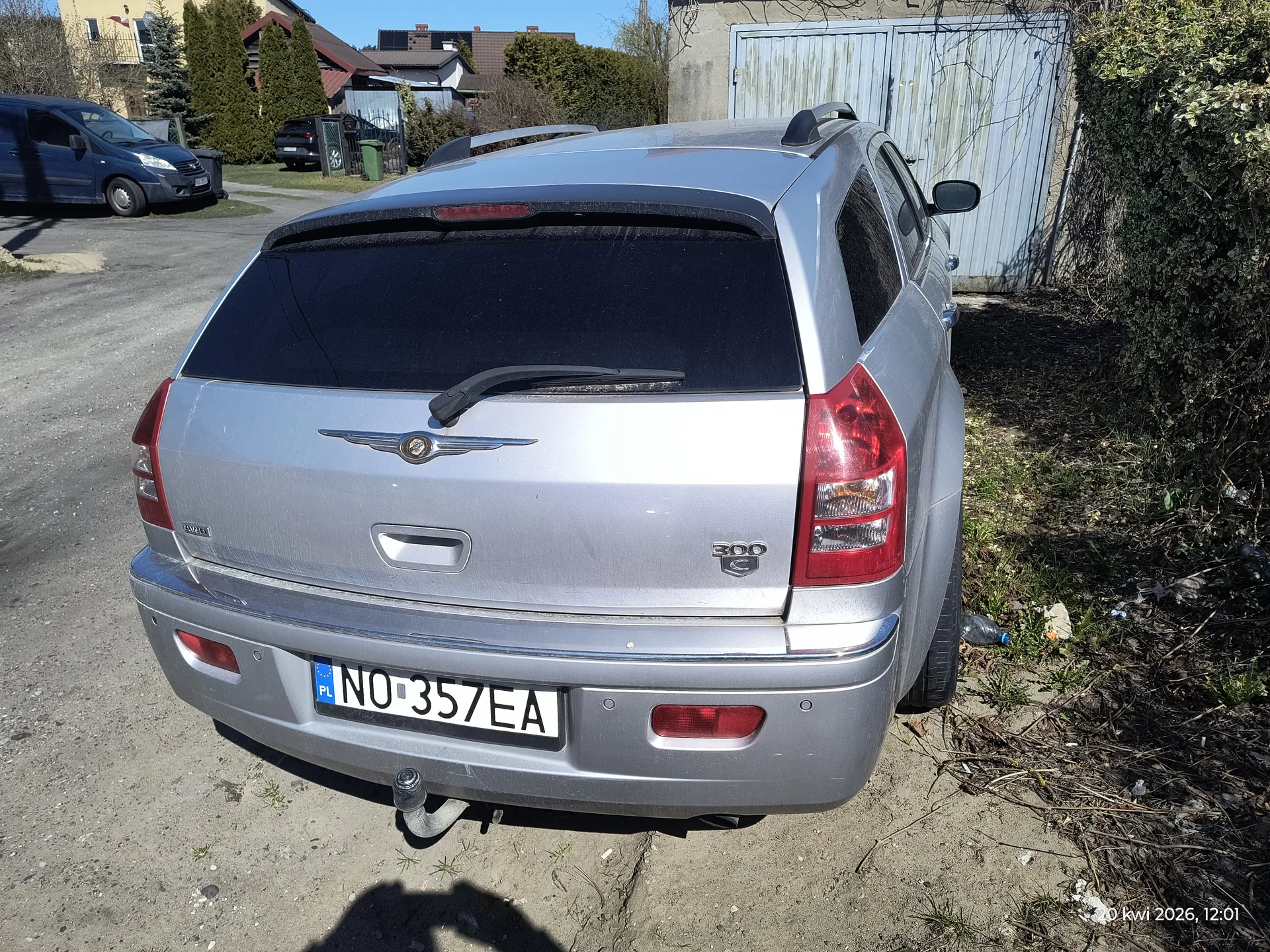 Srebrny samochód osobowy typu kombi marki Chrysler 300C z widocznym logo na bagażniku i napisem "300 C" po prawej stronie. Tablica rejestracyjna PL NO-357EA umieszczona na tylnej części pojazdu. Samochód zaparkowany na żwirowym podjeździe, w tle widoczne są inne pojazdy, domy, krzewy, metalowa brama oraz zielone pojemniki na śmieci. Na prawym dolnym rogu zdjęcia znajduje się znacznik czasu: "20 kwi 2026, 12:01". Na ziemi wokół samochodu widoczne są suche gałęzie i śmieci.