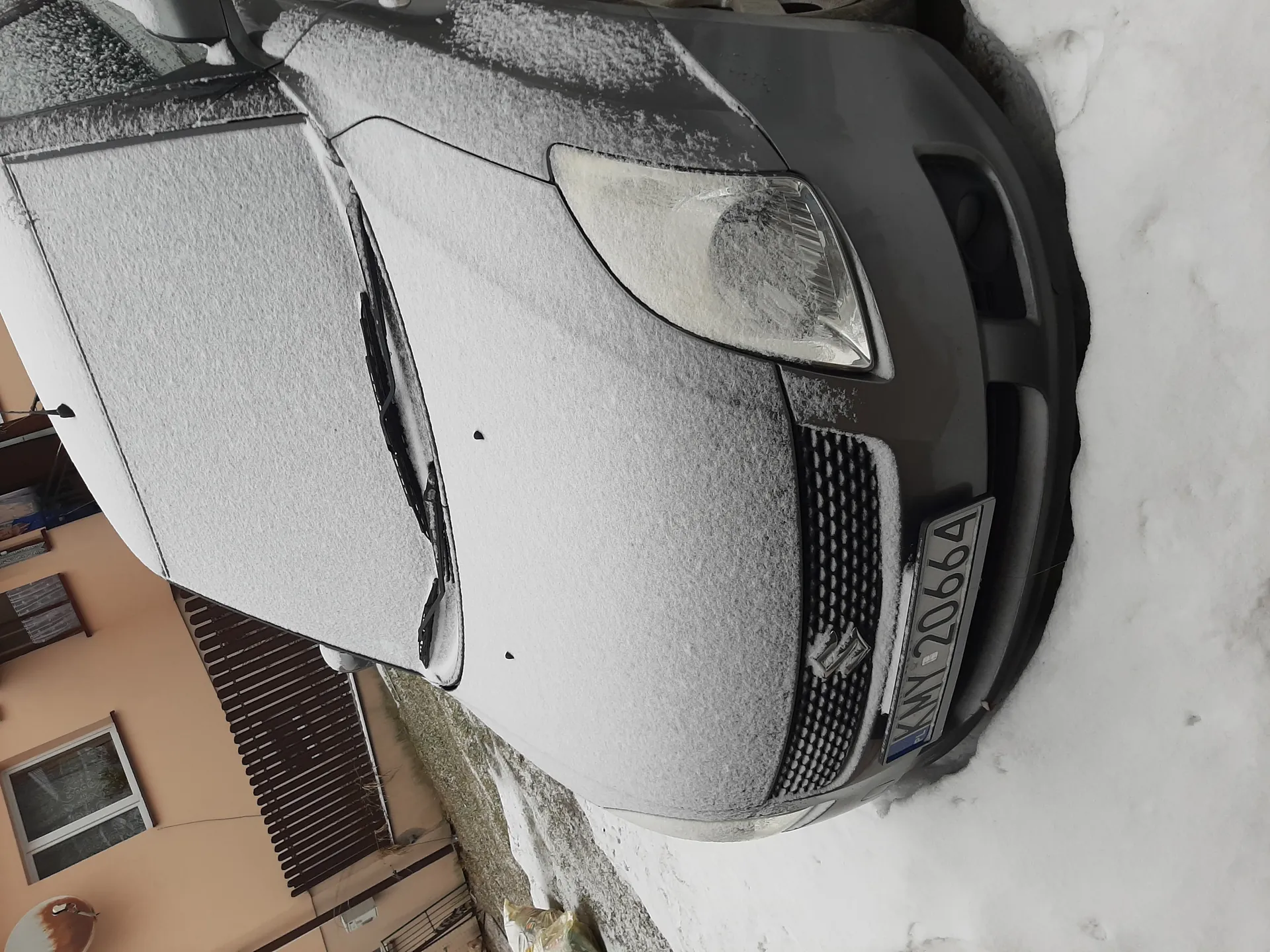 Szary samochód pokryty warstwą śniegu, widoczna tablica rejestracyjna KMY 20664. W tle budynek o beżowej elewacji z oknami, satelitą i bramą. Śnieg otacza pojazd, częściowo zakrywając maskę i reflektory.