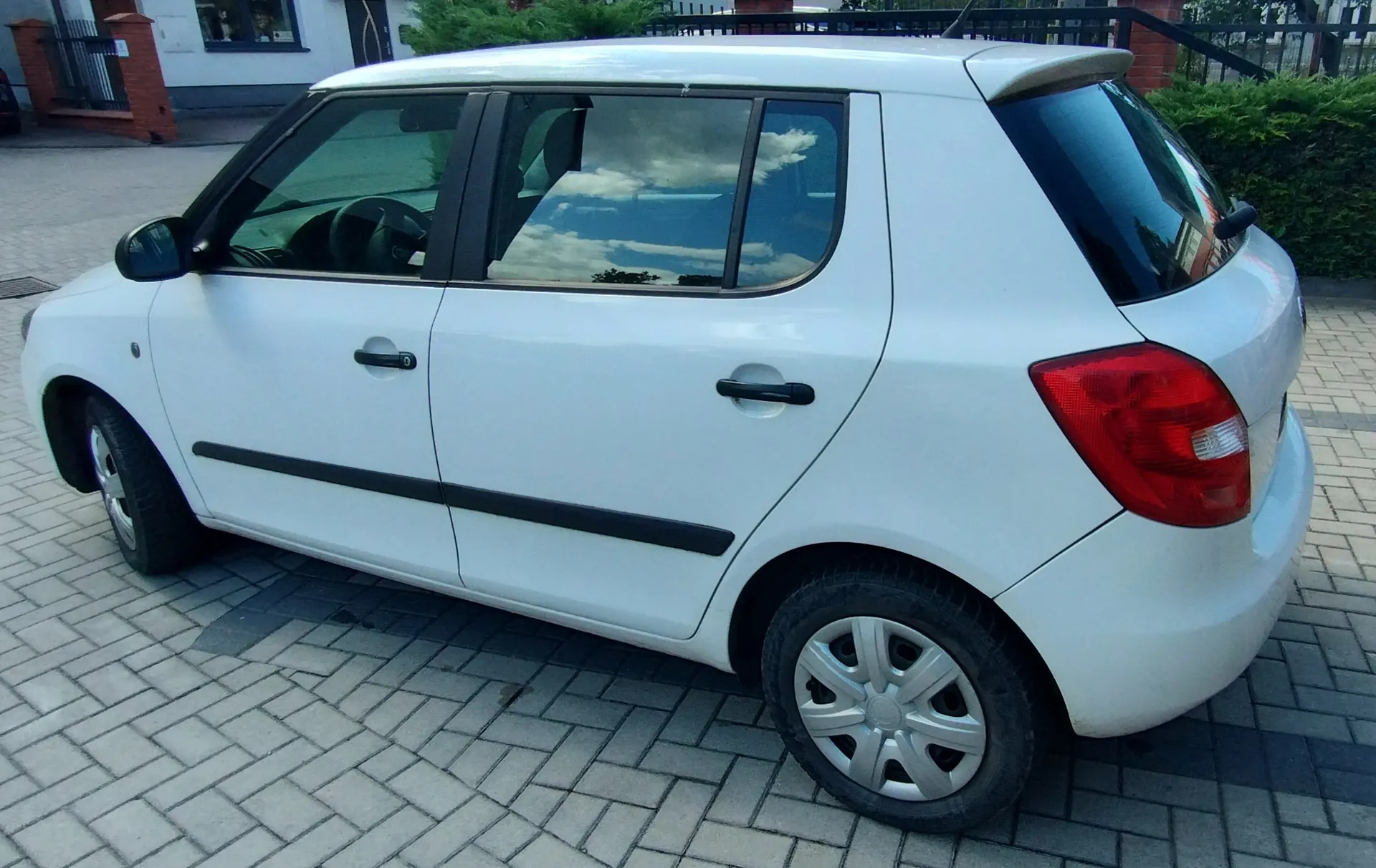 Biały samochód osobowy typu hatchback zaparkowany na kostce brukowej. Pojazd jest pokazany z profilu od strony pasażera, z widocznymi czarnymi listwami ochronnymi na drzwiach i czarnymi klamkami. W tle widoczny jest fragment budynku oraz zielone krzewy.