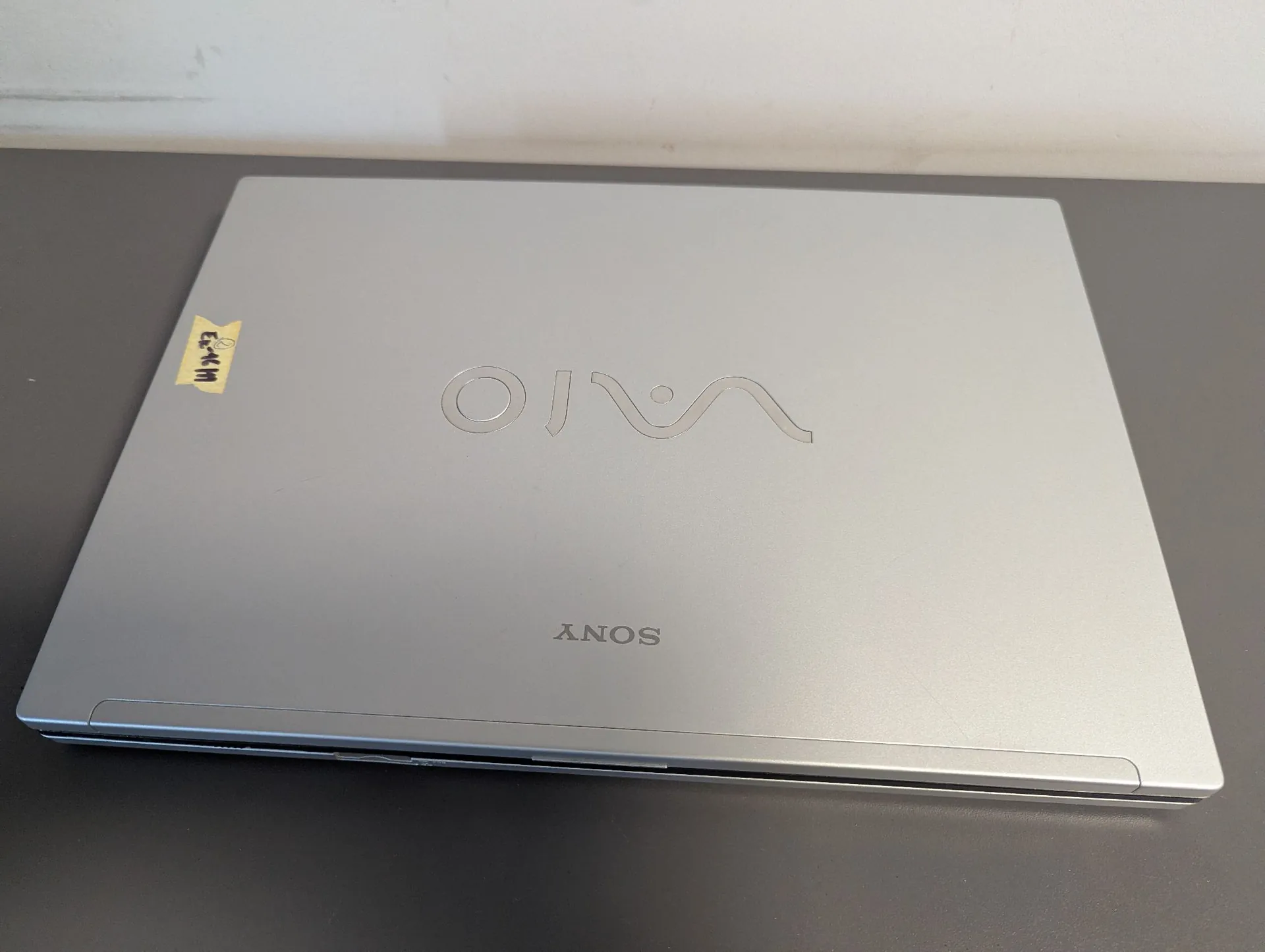 Srebrny laptop marki Sony VAIO zamknięty i leżący na ciemnej powierzchni. Na pokrywie znajduje się duże, wytłoczone logo VAIO oraz napis SONY poniżej. W lewym górnym rogu naklejona jest mała żółta etykieta z ręcznym napisem.