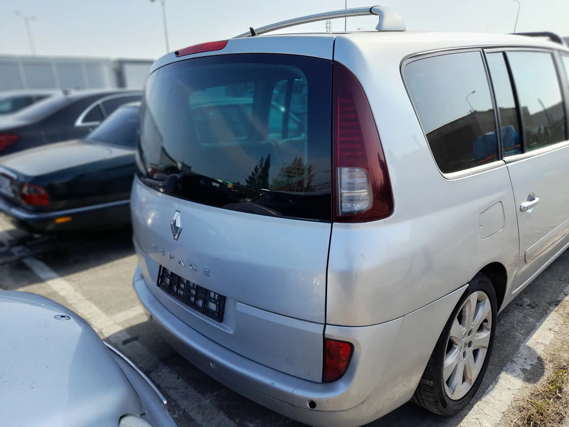 Srebrny samochód Renault Espace widziany od tyłu, z widocznym logo marki i napisem ESPACE na bagażniku. Samochód ma czarny uchwyt na tablicę rejestracyjną bez tablicy, czerwone światła tylnego oraz dachowy bagażnik. W tle znajdują się inne zaparkowane pojazdy, w tym ciemny sedan, na asfaltowym parkingu z białymi liniami. Tło obejmuje budynki i jasne niebo.