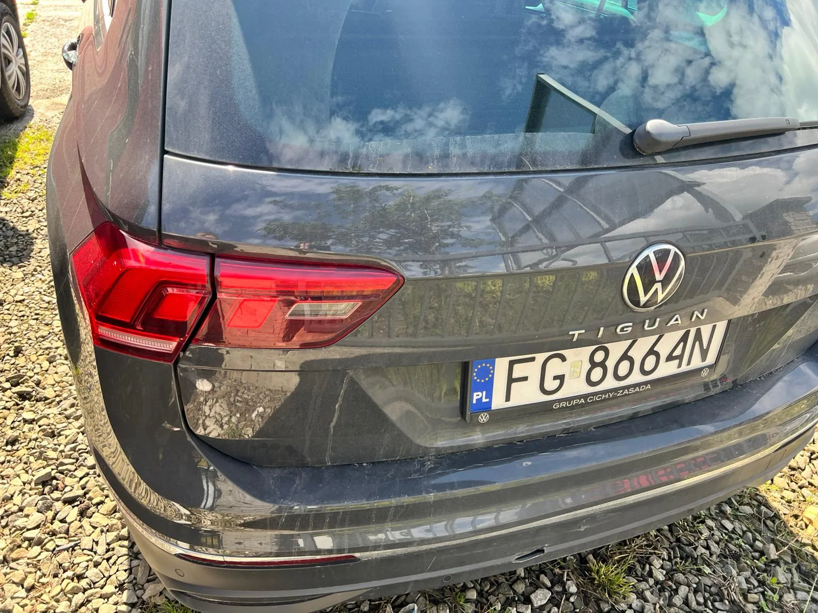 Tylny widok ciemnoszarego samochodu marki Volkswagen Tiguan. Widoczne jest logo marki, napis TIGUAN oraz polska tablica rejestracyjna o numerze FG 8664N. Na ramce tablicy widnieje napis GRUPA CICHY-ZASADA. Samochód stoi na żwirowym podłożu.