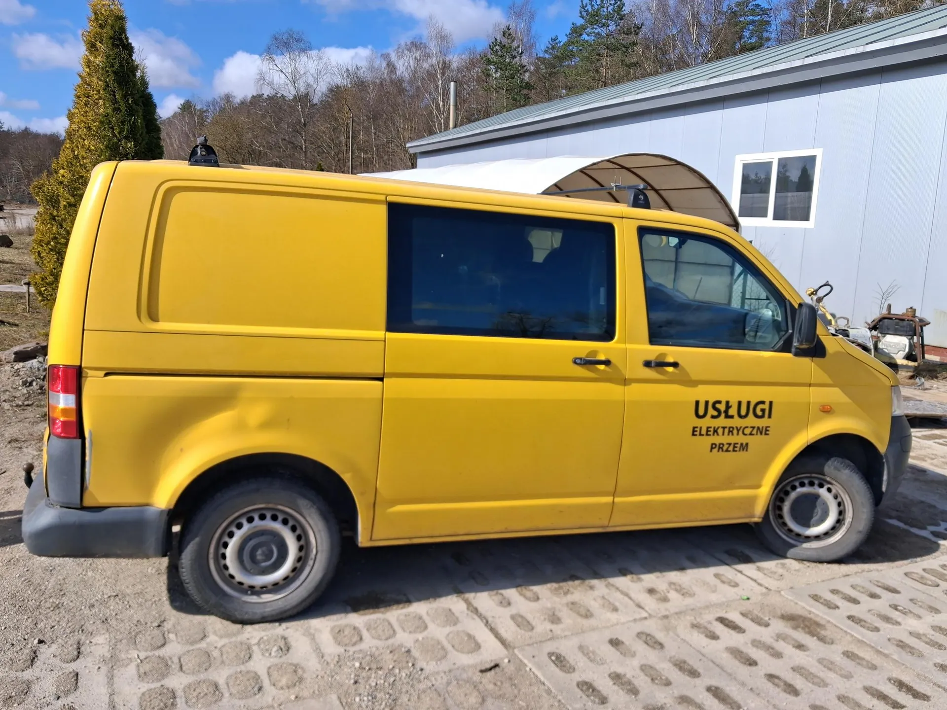 Żółty samochód dostawczy zaparkowany na podwórku. Na boku pojazdu widoczny czarny napis: USŁUGI ELEKTRYCZNE PRZEM. W tle budynek o jasnoszarej elewacji z oknem, drzewa i niebo z chmurami. Samochód ma ciemne szyby, czarne klamki i felgi. Podłoga składa się z kamiennych płyt.
