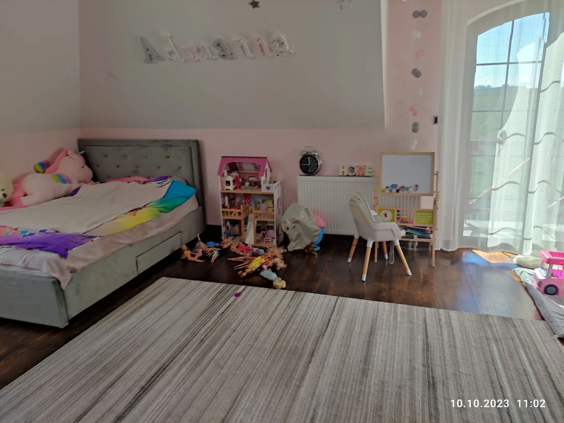 Wnętrze pokoju dziecięcego z łóżkiem z szarym zagłówkiem i kolorową pościelą w tęczowe wzory. Na ścianie nad łóżkiem znajduje się dekoracyjny napis 'Adrianna'. W pokoju widoczny jest różowy domek dla lalek, biały grzejnik, małe białe krzesło oraz sztaluga z rysunkami. Na ciemnej drewnianej podłodze leży szary dywan w prążki oraz rozrzucone zabawki. Po prawej stronie znajduje się okno z białymi firankami, przez które wpada światło. W prawym dolnym rogu widnieje data i godzina: 10.10.2023 11:02.