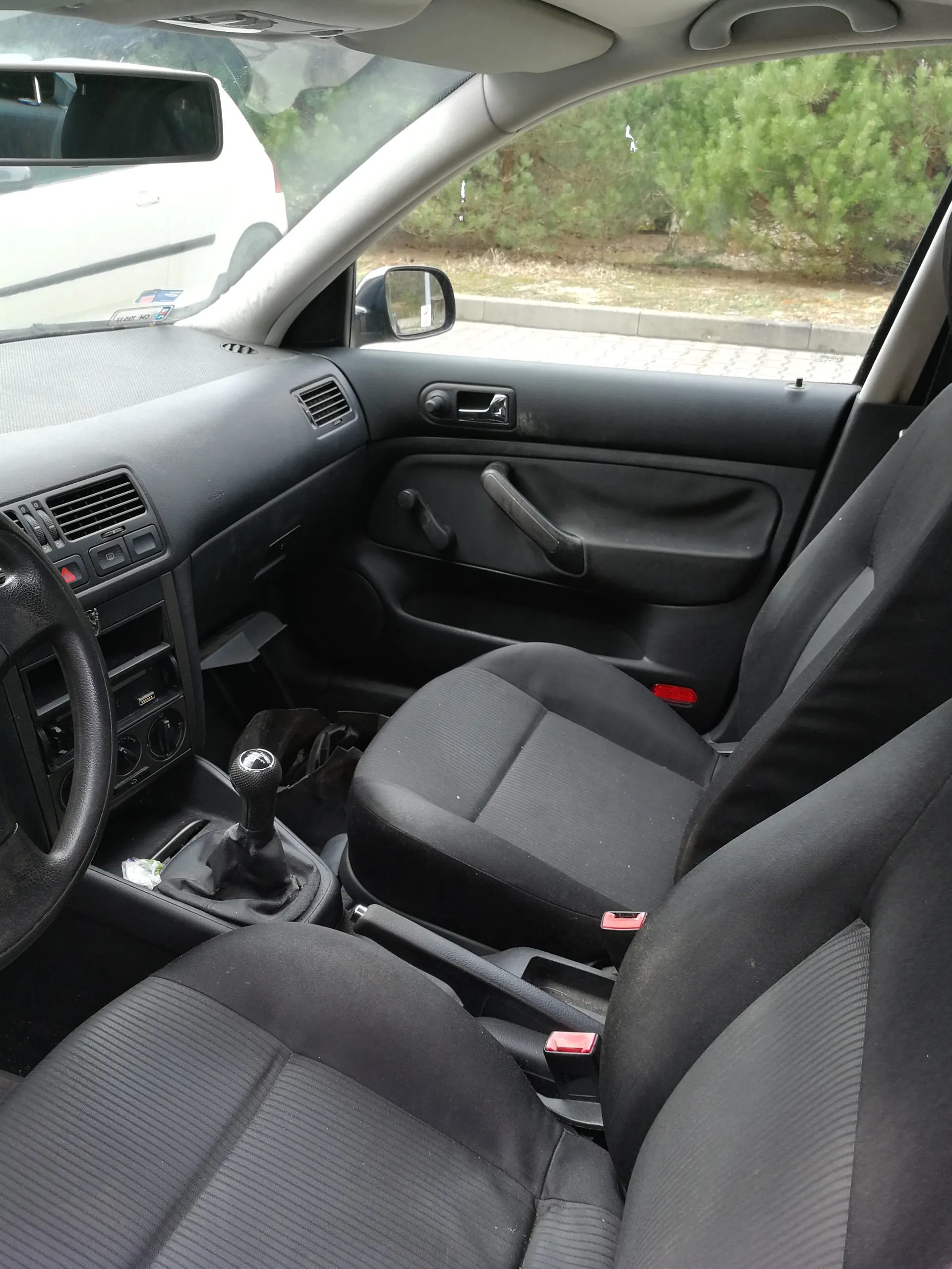 Wnętrze samochodu z widoczną czarną tapicerką siedzeń, kierownicą po lewej stronie, dźwignią zmiany biegów, deska rozdzielcza z przyciskami i wentylacją. W tle przez przednią szybę widać biały samochód oraz zielone drzewa i chodnik. Wnętrze jest czyste, a elementy takie jak lusterko wsteczne i uchwyty drzwiowe są wyraźnie widoczne.