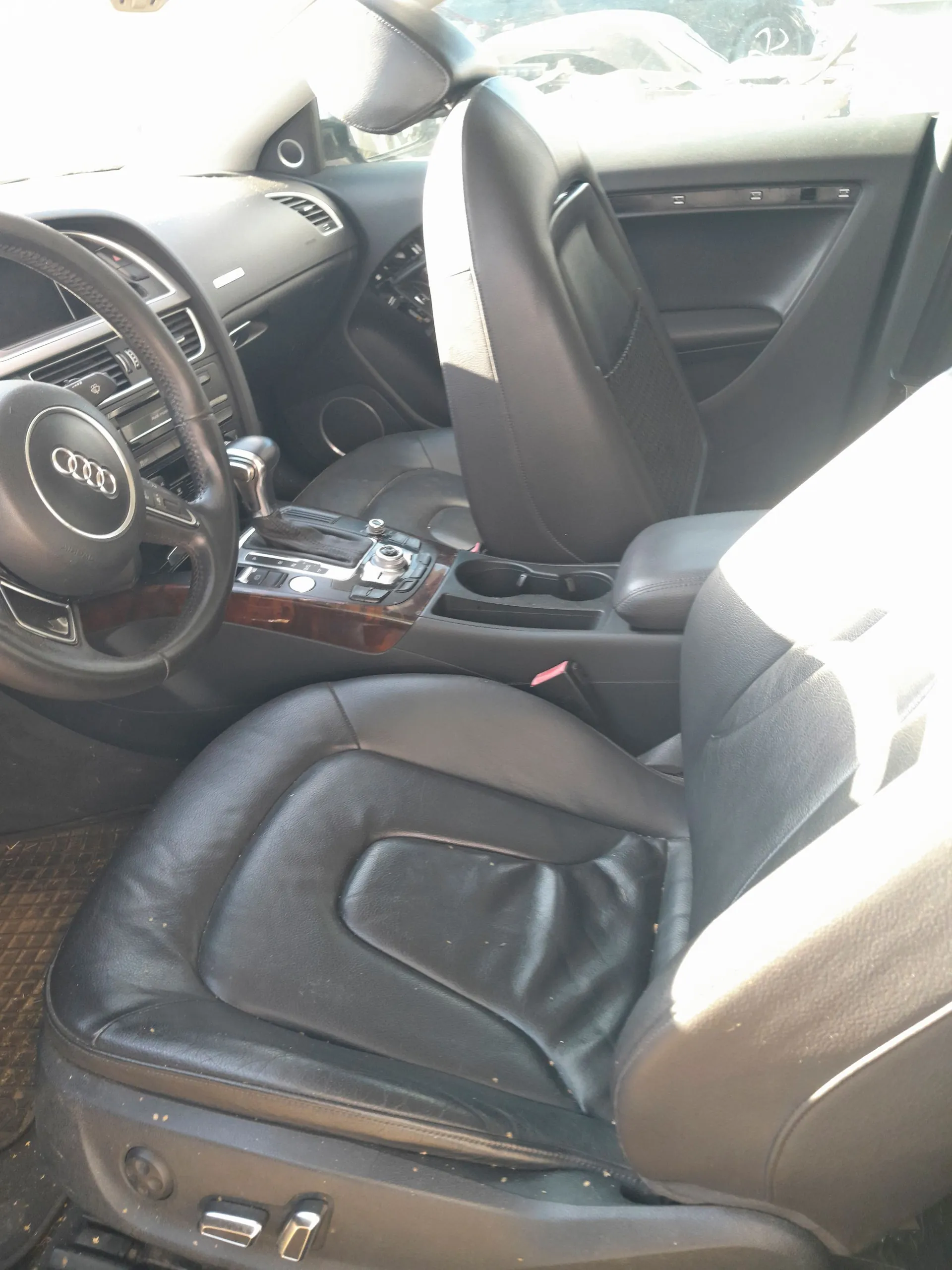 Wnętrze samochodu marki Audi z czarną skórzaną tapicerką. Widoczny jest fotel kierowcy, fotel pasażera, kierownica z logo Audi oraz konsola środkowa z elementami drewnianego wykończenia i przyciskami sterowania. W tle widoczny jest panel drzwi oraz fragment deski rozdzielczej.