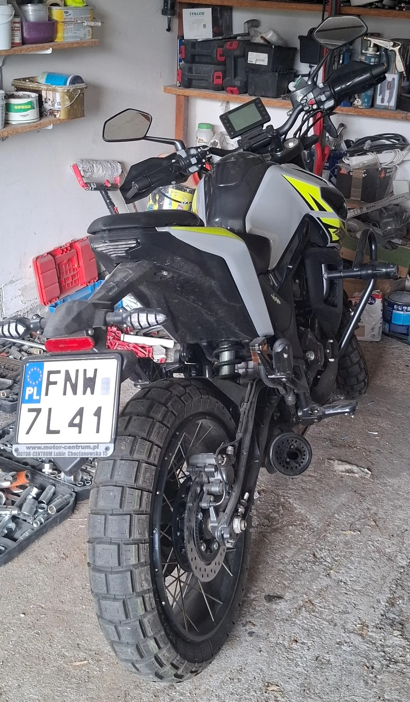 Motocykl w kolorze czarno-szarym z żółtymi akcentami, widok od tyłu, z tablicą rejestracyjną PL FNW 7L41 oraz napisem www.motor-centrum.pl na dole. W tle garaż z półkami zawierającymi narzędzia, pojemniki i pudełka. Podłoga z betonu, widoczne elementy wyposażenia garażowego.