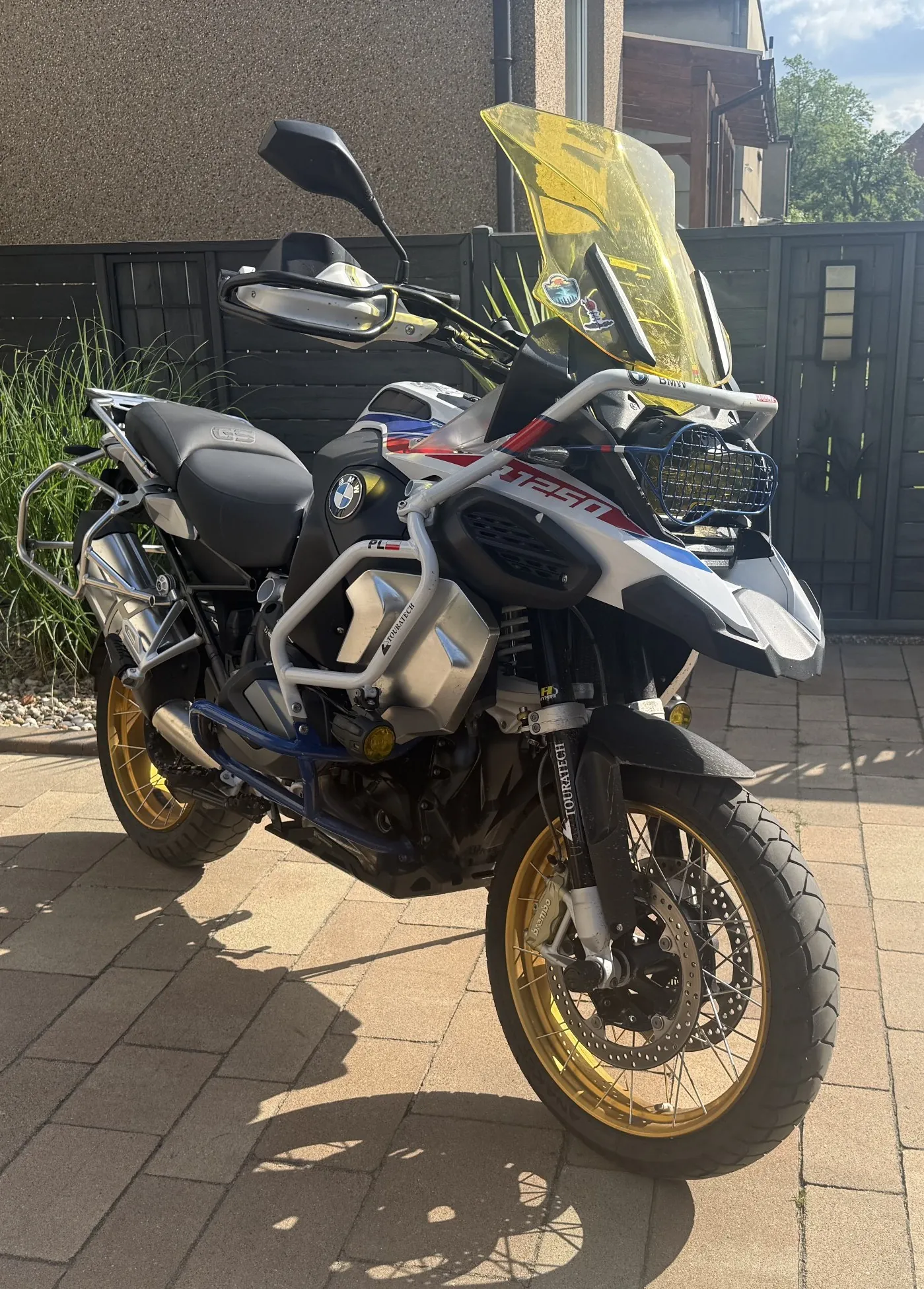 Jasny motocykl BMW R 1250 GS Adventure zaparkowany na kostce brukowej przed domem. Motocykl ma biało-niebiesko-czerwone malowanie, złote felgi, żółtą szybę przednią oraz dodatkowe wyposażenie w postaci srebrnych osłon silnika i bagażnika. Na boku widnieje napis '1250' oraz logo BMW.