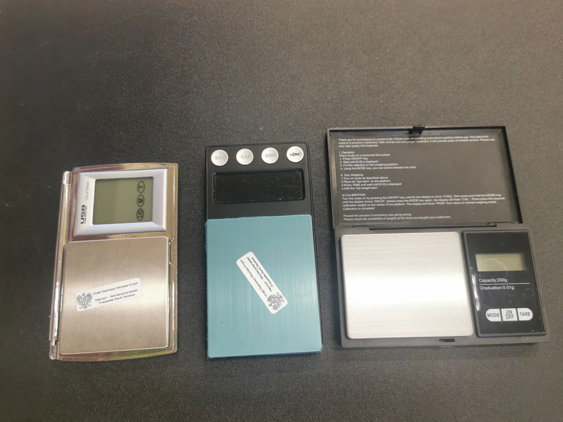 Trzy elektroniczne wagi kuchenne ułożone obok siebie na ciemnym, teksturowanym blacie. Lewa waga jest srebrna z wyświetlaczem LCD i napisem 'USB CALIFORNIA'. Środkowa waga jest błękitna, z naklejoną białą etykietą z kodem QR i tekstem. Prawa waga jest czarna, posiada wyświetlacz LCD oraz trzy przyciski: 'MODE', 'ON OFF' i 'TARE'. Nad czarną wagą znajduje się instrukcja obsługi w języku angielskim, zawierająca sekcje takie jak 'Tares Weighing' oraz 'CALIBRATION', a także informację o pojemności 'Capacity: 200g' i dokładności 'Graduation: 0.01g'.