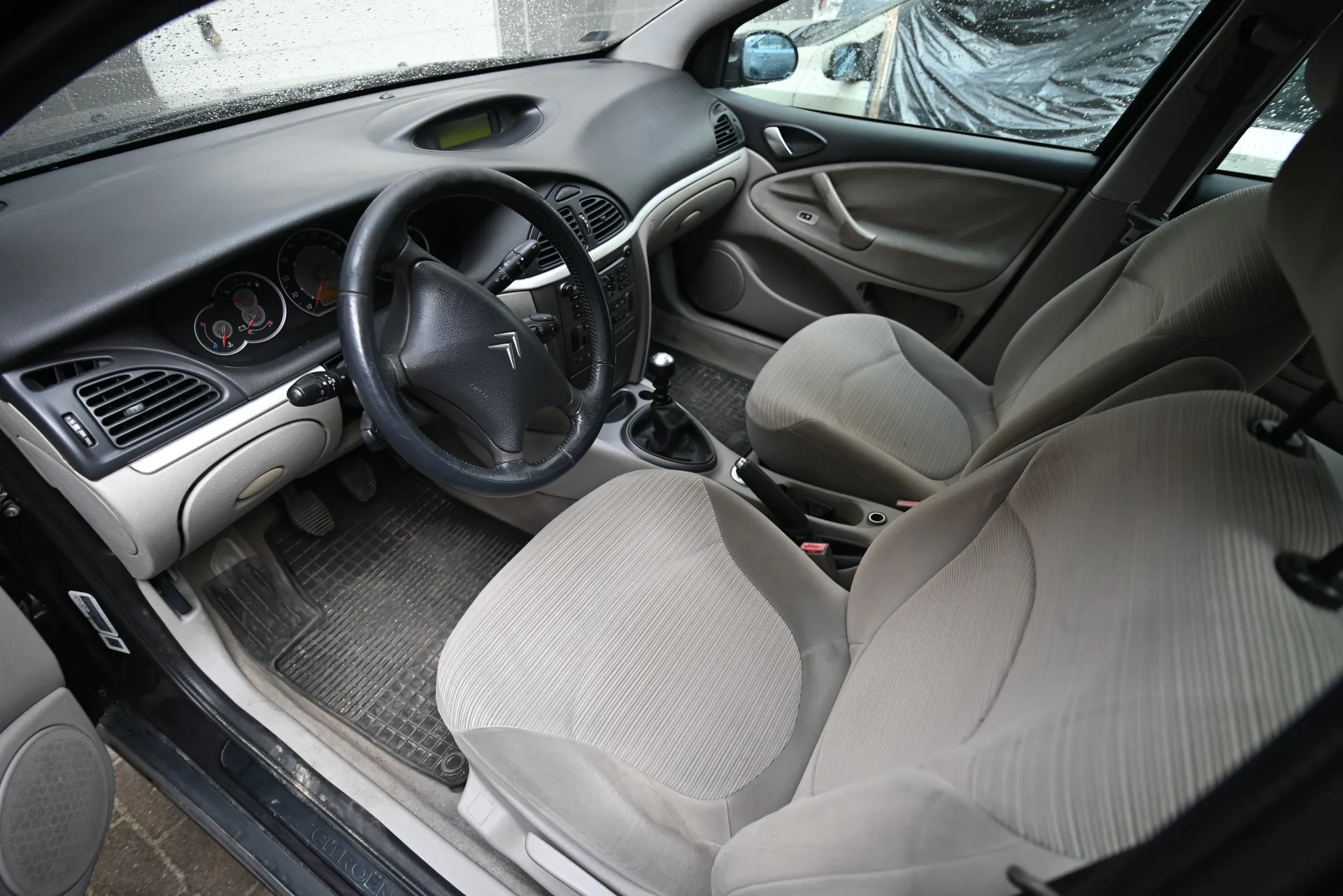 Wnętrze samochodu Citroën z widoczną kierownicą z logo marki, szarym deskiem rozdzielczym z dwoma okrągłymi wskaźnikami, szarymi siedzeniami z pionową fakturą, dźwignią zmiany biegów i czarnymi dywanikami. Widoczne są otwarte drzwi, deszcz na przedniej szybie oraz budynki zasłonięte folią w tle. Na progu drzwi widoczny napis 'CITROËN'.