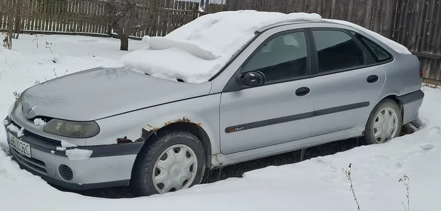 Srebrny samochód osobowy typu sedan zaparkowany na śniegu. Na dachu i masce widoczna warstwa śniegu. Przednie koło po lewej stronie z widoczną rdzą wokół błotnika. Tablica rejestracyjna z napisem AB0032C. W tle drewniany płot i drzewo bez liści. Śnieg pokrywa całą powierzchnię ziemi.