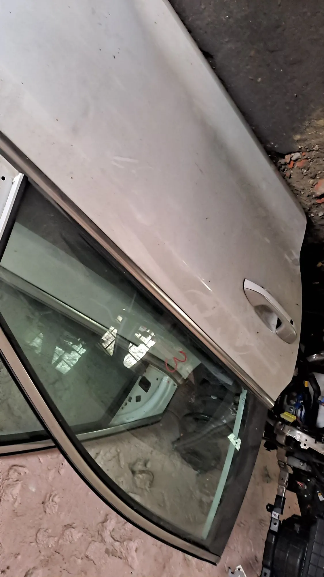 Zbliżenie na białe drzwi samochodu z szybą. Samochód jest brudny, a drzwi znajdują się w nienaturalnej pozycji, prawdopodobnie w warsztacie lub na złomowisku. W dolnej części widoczne są elementy mechaniczne i klamka drzwi.