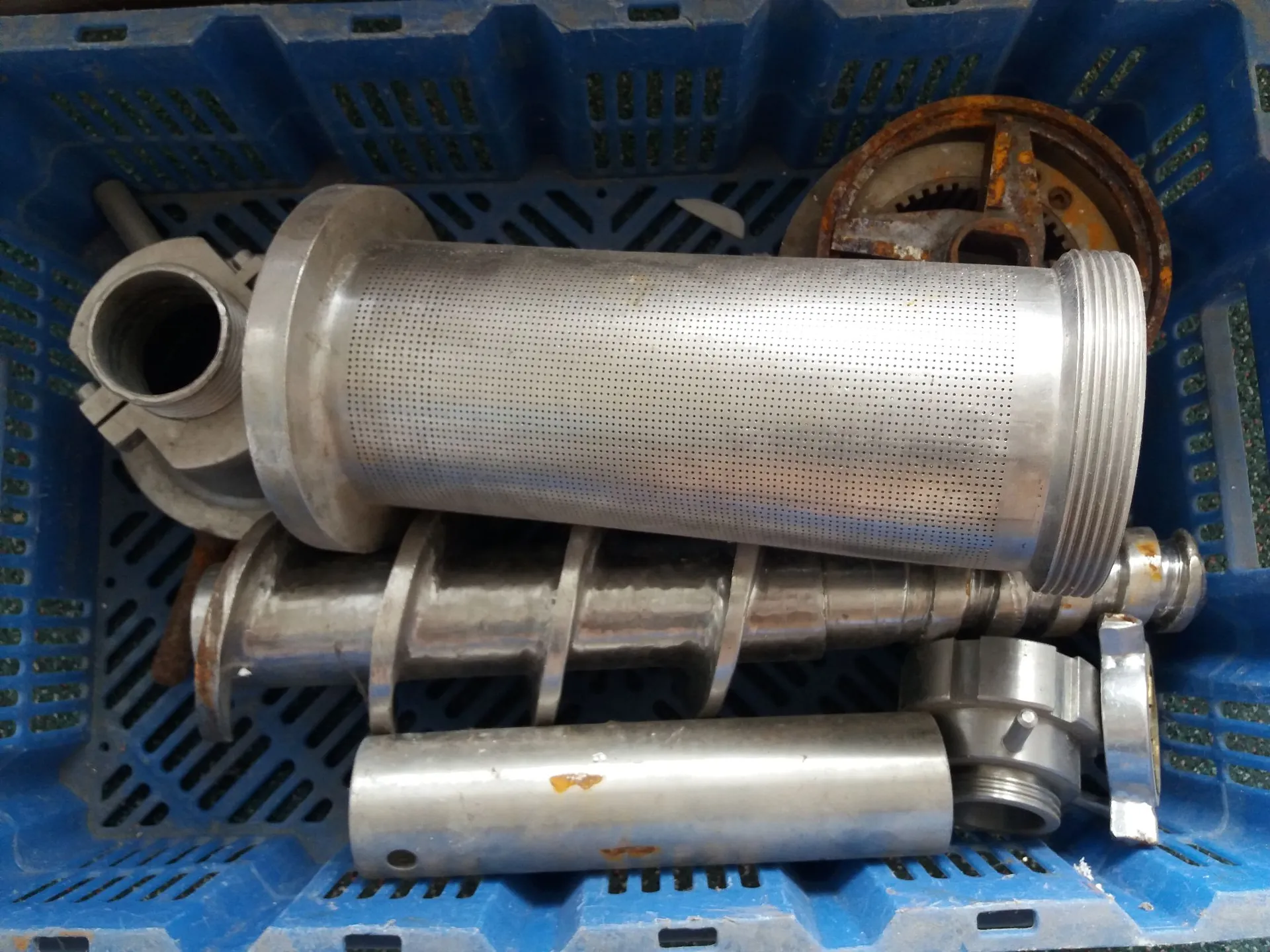 Niebieski plastikowy kosz z dziurkami, w którym znajdują się metalowe części maszyn. Widoczny jest duży, cylindryczny filtr z perforowaną powierzchnią, metalowy wałek z nacięciami w kształcie spirali (ślimak), mniejszy gładki cylinder oraz rdzawa, okrągła tarcza zębata.