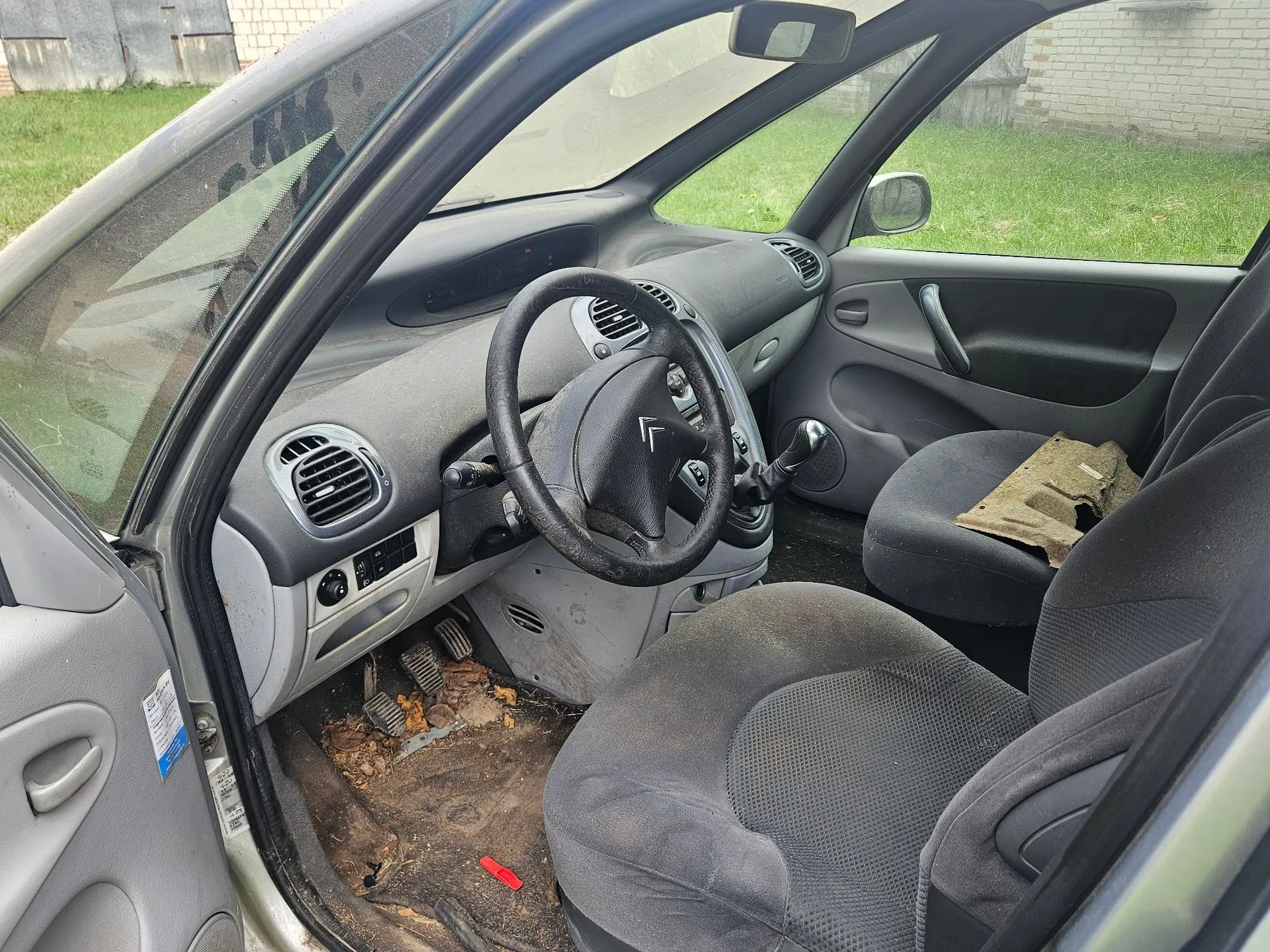 Wnętrze samochodu z widokiem na deskę rozdzielczą, kierownicę z logo Citroëna oraz przednie fotele z ciemnoszarej tkaniny. Podłoga po stronie kierowcy jest brudna, z widocznymi resztkami liści i ziemi. Na siedzeniu pasażera leży kawałek brązowego materiału lub tektury.