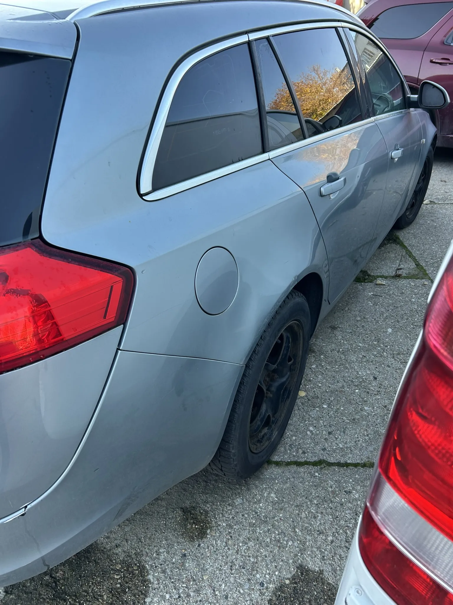 Srebrny samochód typu kombi zaparkowany na betonowym placu parkingowym. Widoczne są czerwone światła tylne oraz okrągła pokrywa zbiornika paliwa po prawej stronie. W tle częściowo widoczne są inne pojazdy, w tym czerwony samochód. Na asfalcie widoczne są plamy i zielone naloty między płytami.