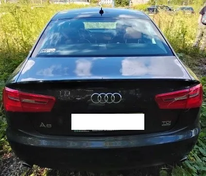 Tylny widok czarnego samochodu marki Audi A6. Na klapie bagażnika widoczne jest logo Audi (cztery splecione pierścienie), oznaczenie modelu 'A6' po lewej stronie oraz oznaczenie silnika 'TDI quattro' po prawej stronie. Tablica rejestracyjna jest zakryta białym prostokątem. Samochód stoi na trawiastym terenie.