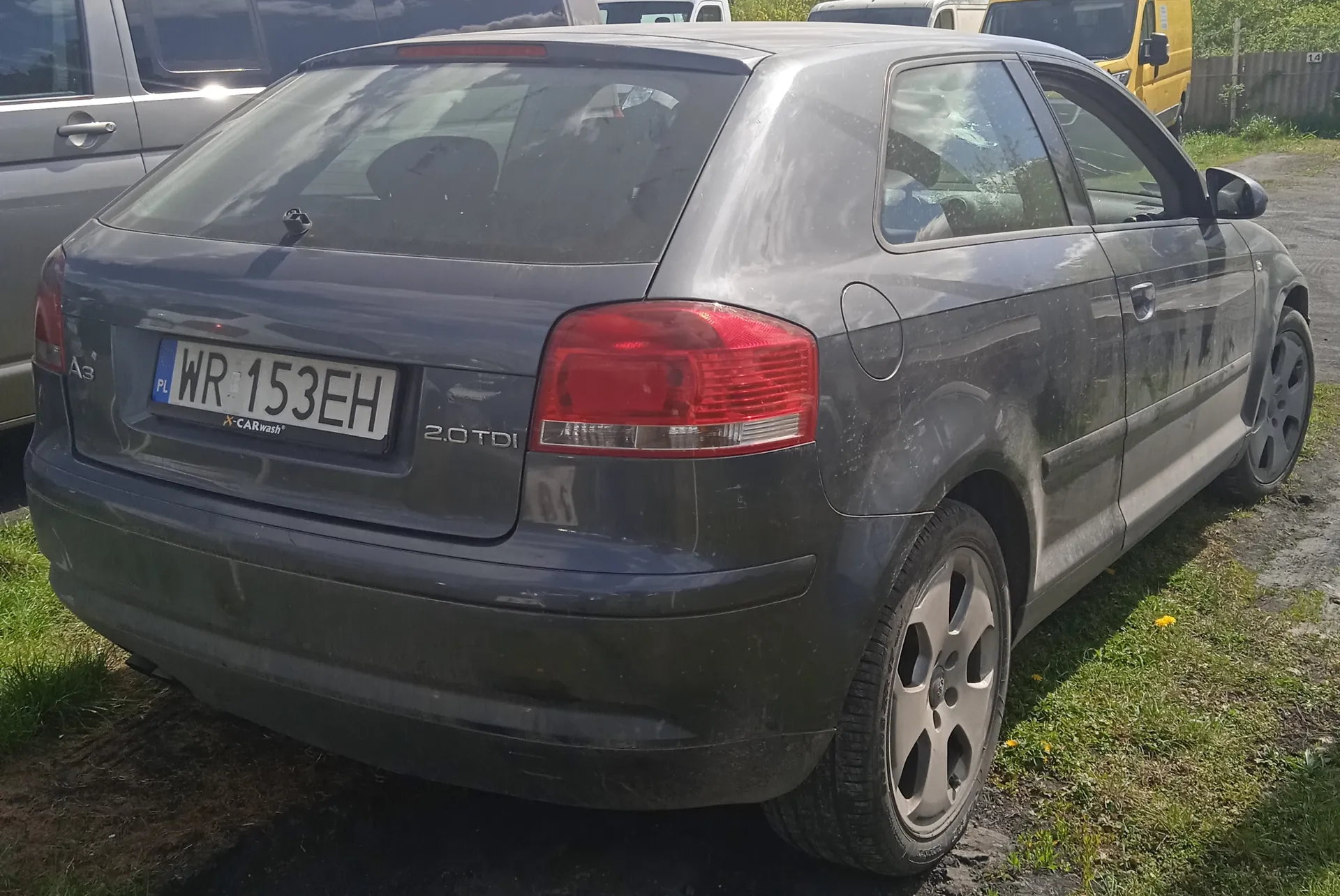 Zdjęcie przedstawia tylną część szarego samochodu Audi A3 z silnikiem 2.0 TDI. Na tablicy rejestracyjnej widoczny jest numer WR 153EH z oznaczeniem PL oraz naklejka CARWASH. Samochód jest zabrudzony, zaparkowany na trawie obok drogi. W tle znajdują się inne pojazdy: srebrny van po lewej stronie i żółty van w oddali. Na trawie widoczne są żółte kwiaty.