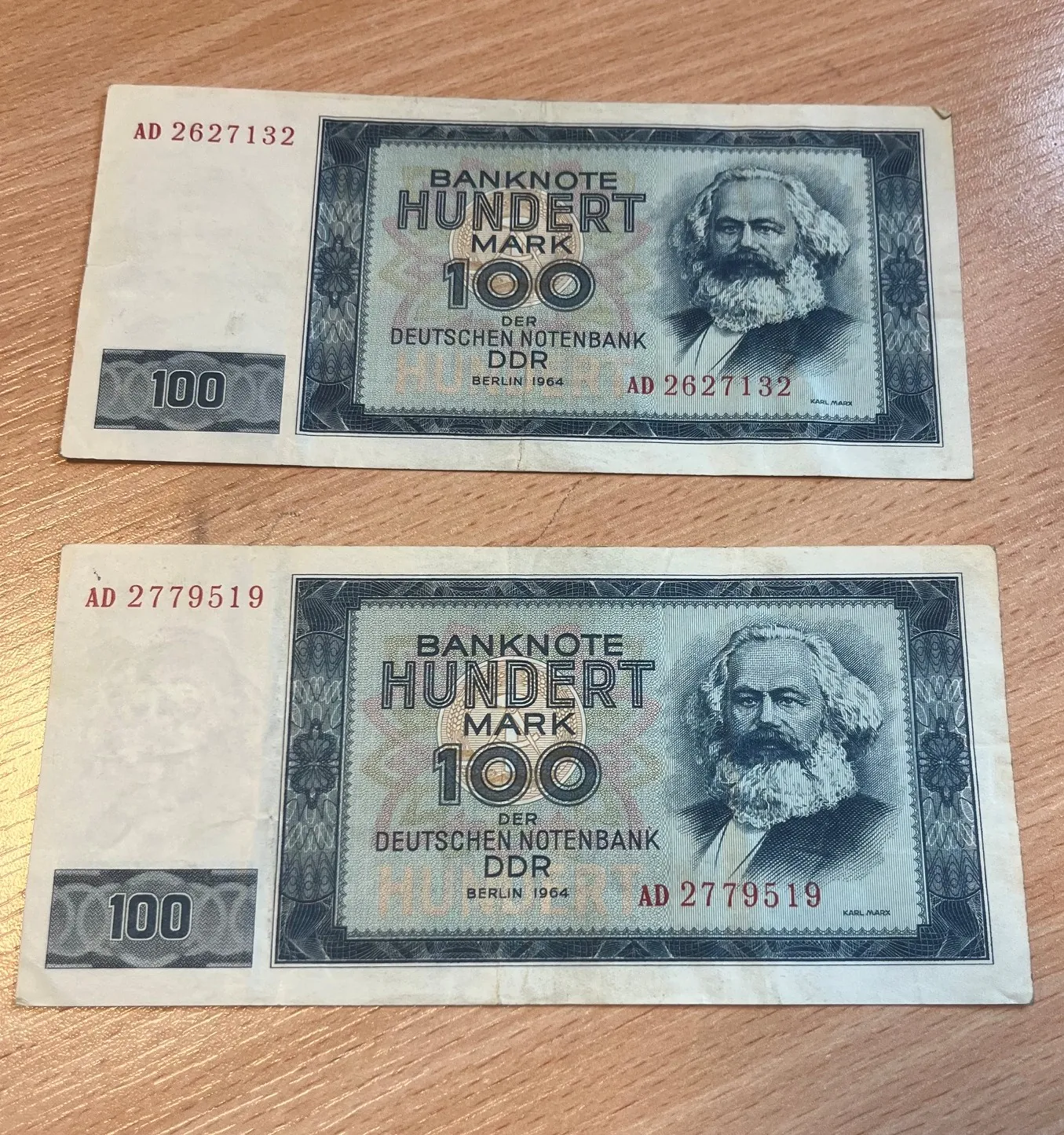 Dwa banknoty o nominale 100 marek z Niemieckiej Republiki Demokratycznej (DDR) z 1964 roku, ułożone jeden nad drugim na drewnianym blacie. Na każdym banknocie widnieje portret Karola Marksa oraz napisy: 'BANKNOTE HUNDERT MARK 100 DER DEUTSCHEN NOTENBANK DDR BERLIN 1964'. Górny banknot ma numer seryjny AD 2627132, a dolny AD 2779519.