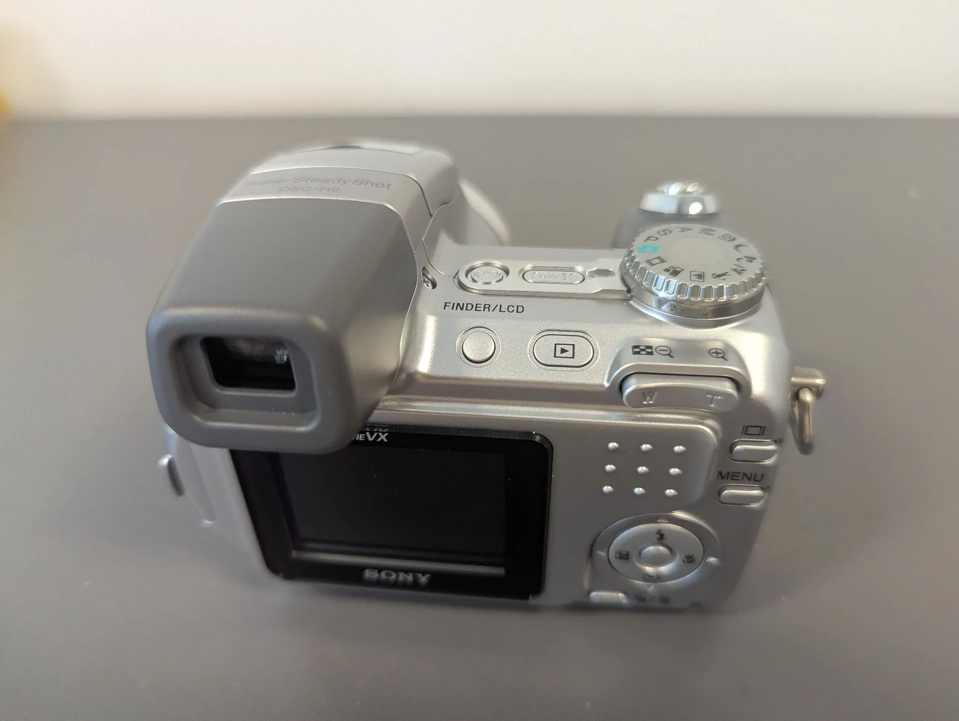Srebrny aparat cyfrowy Sony Cyber-shot widziany od tyłu. Widoczny jest wizjer, ekran LCD, przyciski sterujące, w tym przycisk MENU oraz pokrętło trybów. Na obudowie znajduje się napis 'Super SteadyShot DSC-H2' oraz 'FINDER/LCD'.