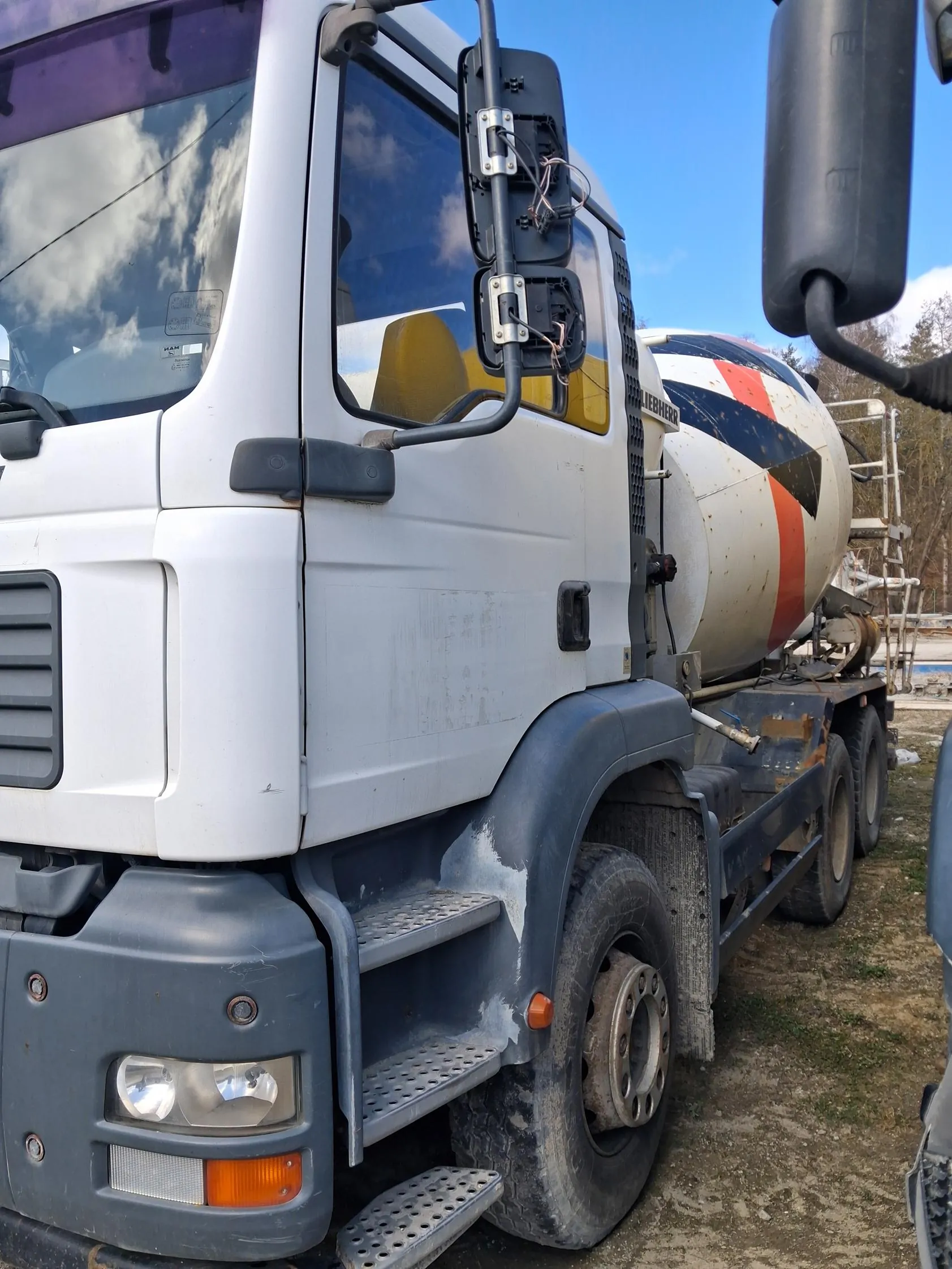 Biały ciężarówka z betoniarką marki Liebherr, z widocznym czerwono-niebieskim pasem na zbiorniku. Pojazd stoi na brudnej, trawiastej powierzchni. W tle drzewa, konstrukcje budowlane i błękitne niebo z chmurami. Widoczne lusterka, kierunkowskazy oraz schodki prowadzące do kabiny. Tekst 'Liebherr' na boku kabiny jest czytelny.