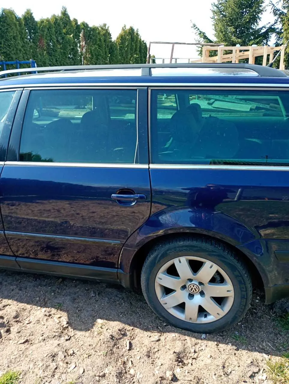 Boczny widok ciemnoniebieskiego samochodu typu kombi marki Volkswagen zaparkowanego na podłożu z ziemi i drobnych kamieni. Widoczne są czarne listwy okienne, dachówka z przenośnikiem, przyciemniane szyby oraz felga z logo VW. W tle drzewa i drewniana konstrukcja. Opona przedniego koła ma widoczny napis 'TOYO'.
