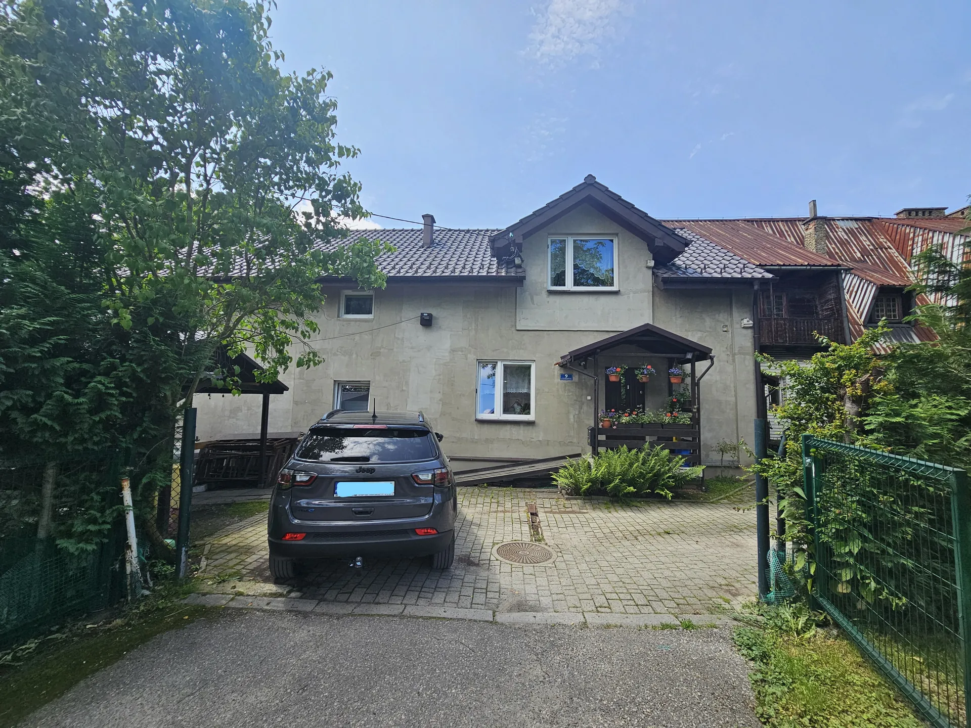 Dwupiętrowy dom o beżowej elewacji z dachem krytym dachówką i blachą. Przed domem stoi szary samochód typu SUV z tyłem skierowanym do obserwatora. Dom posiada kilka okien, w tym okno na piętrze z widocznymi kwiatami na parapecie oraz daszek nad wejściem z wiszącymi doniczkami. Po obu stronach domu rosną drzewa i krzewy, a przed nim znajduje się brukowana podjazdowa ścieżka. Po lewej stronie widoczny jest zielony płot, a po prawej zielony metalowy ogrodzenie. Niebo jest jasne z kilkoma chmurami.