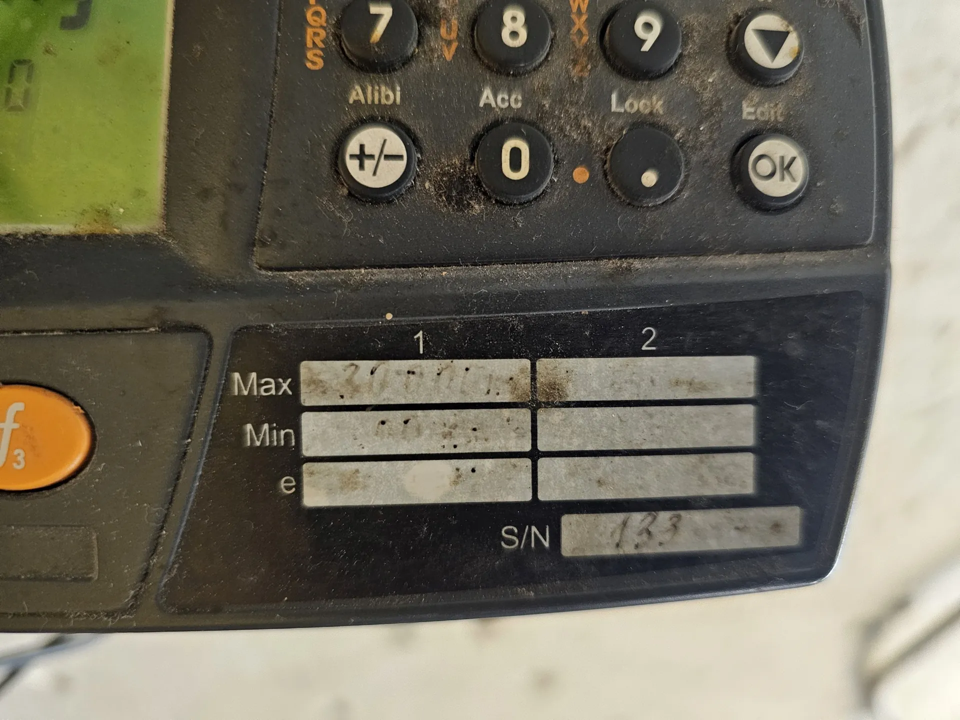 Zbliżenie na czarny panel sterowania urządzenia elektronicznego, prawdopodobnie wagi przemysłowej. W górnej części znajdują się gumowe przyciski z cyframi 7, 8, 9, 0 oraz funkcjami: Alibi, Acc, Lock, Edit, +/- i OK. Poniżej znajduje się srebrna tabliczka znamionowa z polami Max, Min oraz e, które są częściowo starte. Na dole tabliczki widnieje napis S/N oraz numer seryjny zaczynający się od cyfr 133. Urządzenie jest zakurzone.