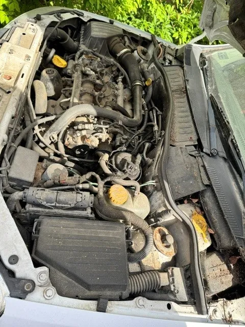 Widok z góry na zakurzoną komorę silnika w samochodzie z otwartą maską. Widoczne są elementy mechaniczne, przewody, czarne plastikowe osłony oraz dwa żółte korki zbiorników płynów.