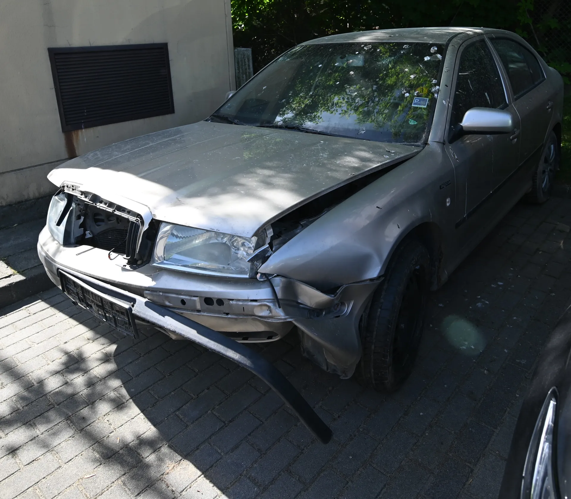Uszkodzony srebrny samochód osobowy z poważnymi uszkodzeniami przodu, w tym zdeformowaną maską, uszkodzonym zderzakiem i brakującym lewym reflektorem. Samochód zaparkowany na brukowanej powierzchni obok budynku z ciemnym wentylatorem na ścianie. W tle zielone drzewa i krzewy. Na przedniej szybie widoczne są ślady uszkodzeń oraz mała naklejka.