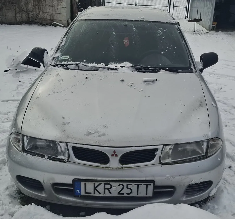 Srebrny samochód marki Mitsubishi pokryty śniegiem, widok z przodu. Na masce widoczny czerwony emblemat marki. Tablica rejestracyjna z napisem LKR 25TT oraz oznaczeniem PL. Przednią szybę i maskę częściowo przykrywa śnieg. W tle budynek, ogrodzenie i śnieżna powierzchnia ziemi.