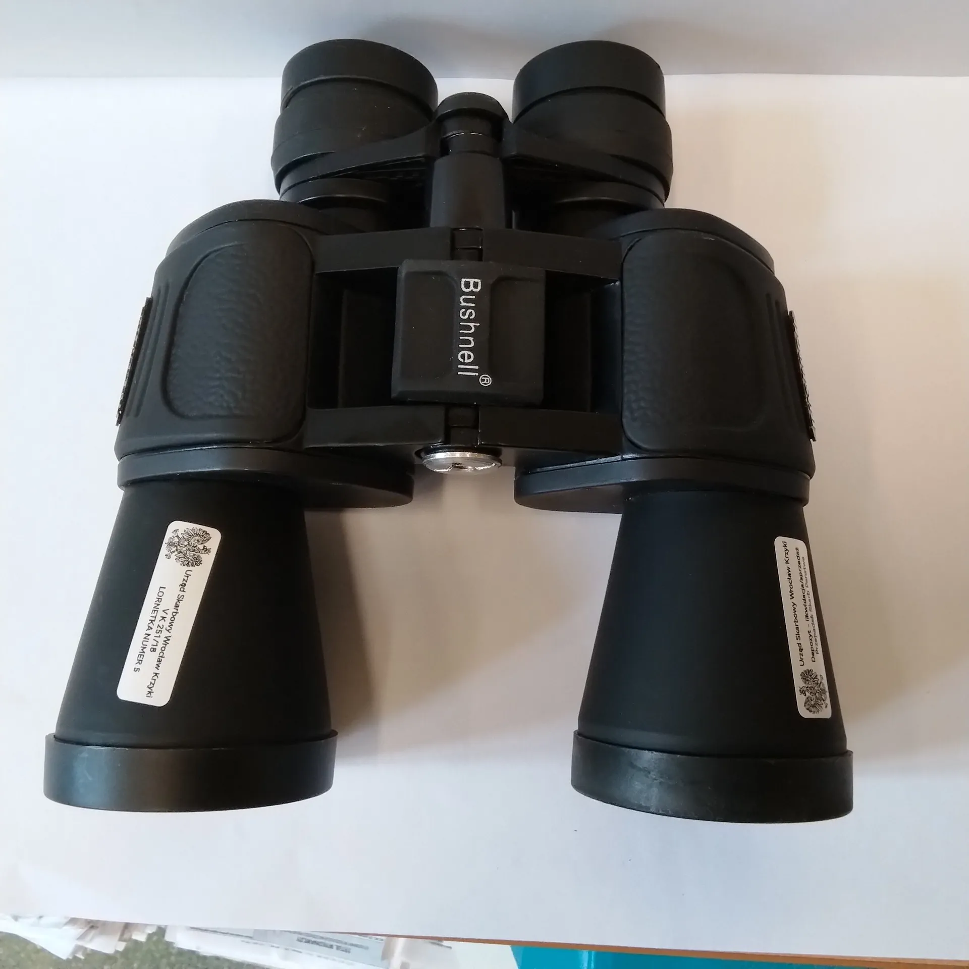 Czarna lornetka marki Bushnell ułożona na białym tle. Na środkowej części urządzenia widnieje pionowy napis 'Bushnell®'. Na obu obiektywach naklejone są białe etykiety z tekstem, z których jedna zawiera napis 'LORNETKA NUMER 5'.