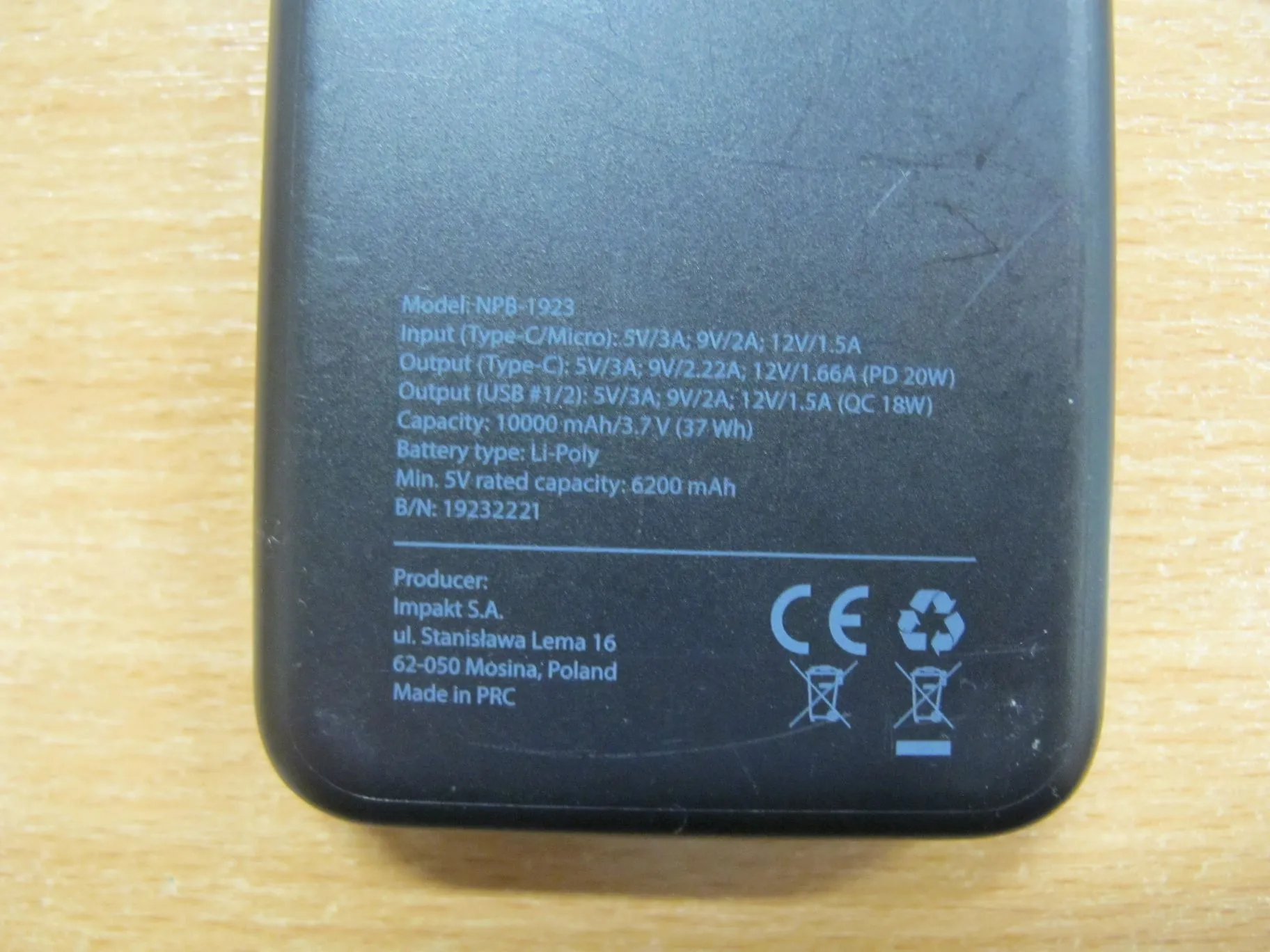 Zbliżenie na tylną część czarnego powerbanku z białymi napisami technicznymi. Tekst zawiera informacje: Model: NPB-1923, Input (Type-C/Micro): 5V/3A; 9V/2A; 12V/1.5A, Output (Type-C): 5V/3A; 9V/2.22A; 12V/1.66A (PD 20W), Output (USB #1/2): 5V/3A; 9V/2A; 12V/1.5A (QC 18W), Capacity: 10000 mAh/3.7 V (37 Wh), Battery type: Li-Poly, Min. 5V rated capacity: 6200 mAh, B/N: 19232221. Poniżej widnieją dane producenta: Producer: Impakt S.A., ul. Stanisława Lema 16, 62-050 Mosina; Poland, Made in PRC. Po prawej stronie znajdują się symbole CE oraz znaki recyklingu.