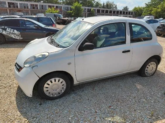 Biały, trzydrzwiowy samochód osobowy zaparkowany na żwirowym placu. Auto jest widoczne z profilu od strony kierowcy. W tle znajdują się inne zaparkowane samochody oraz długi, wielopiętrowy budynek o charakterze przemysłowym lub szkolnym z licznymi oknami.