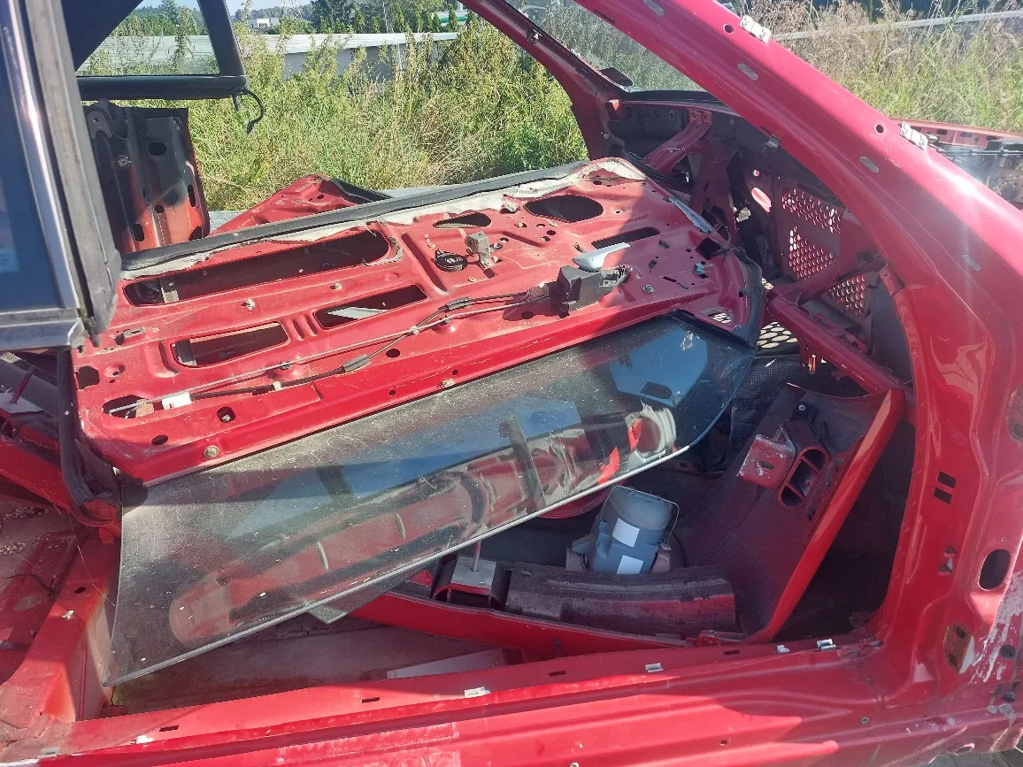 Wnętrze czerwonego samochodu w stanie rozbiórki lub renowacji. Widoczna jest metalowa konstrukcja deski rozdzielczej, elementy nadwozia oraz duża, przezroczysta szyba przednia leżąca ukośnie wewnątrz kabiny. W tle widać zieloną roślinność i fragment ogrodzenia.