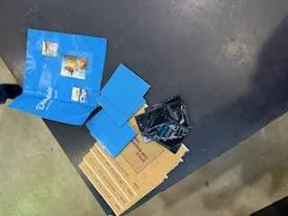 Niebieskie teczki lub koperty z widocznymi etykietami lub zdjęciami, czarna torba plastikowa oraz kawałki kartonu ułożone na ciemnej powierzchni.