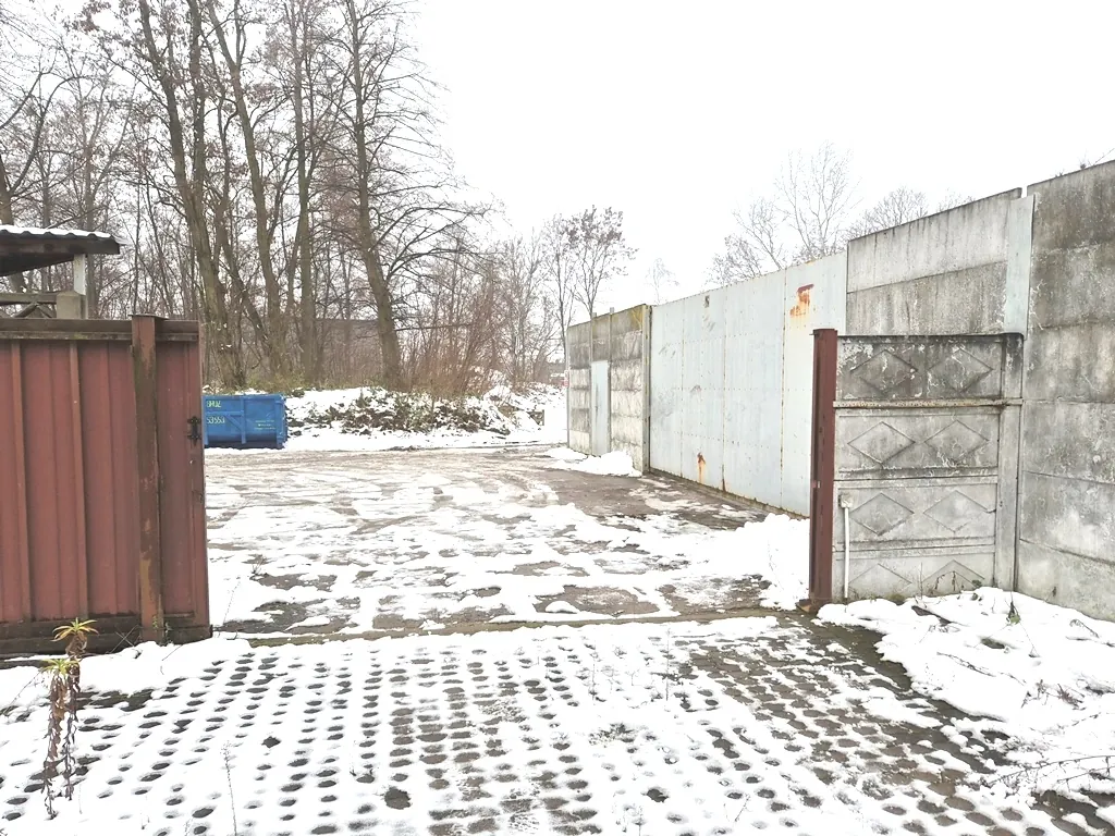 Zimowy widok na utwardzony plac pokryty cienką warstwą śniegu. Po lewej stronie znajduje się brązowy drewniany płot, a po prawej wysoki, betonowy mur z panelami. W tle widać niebieski kontener na odpady oraz gęstą grupę bezlistnych drzew na tle jasnego, zachmurzonego nieba.