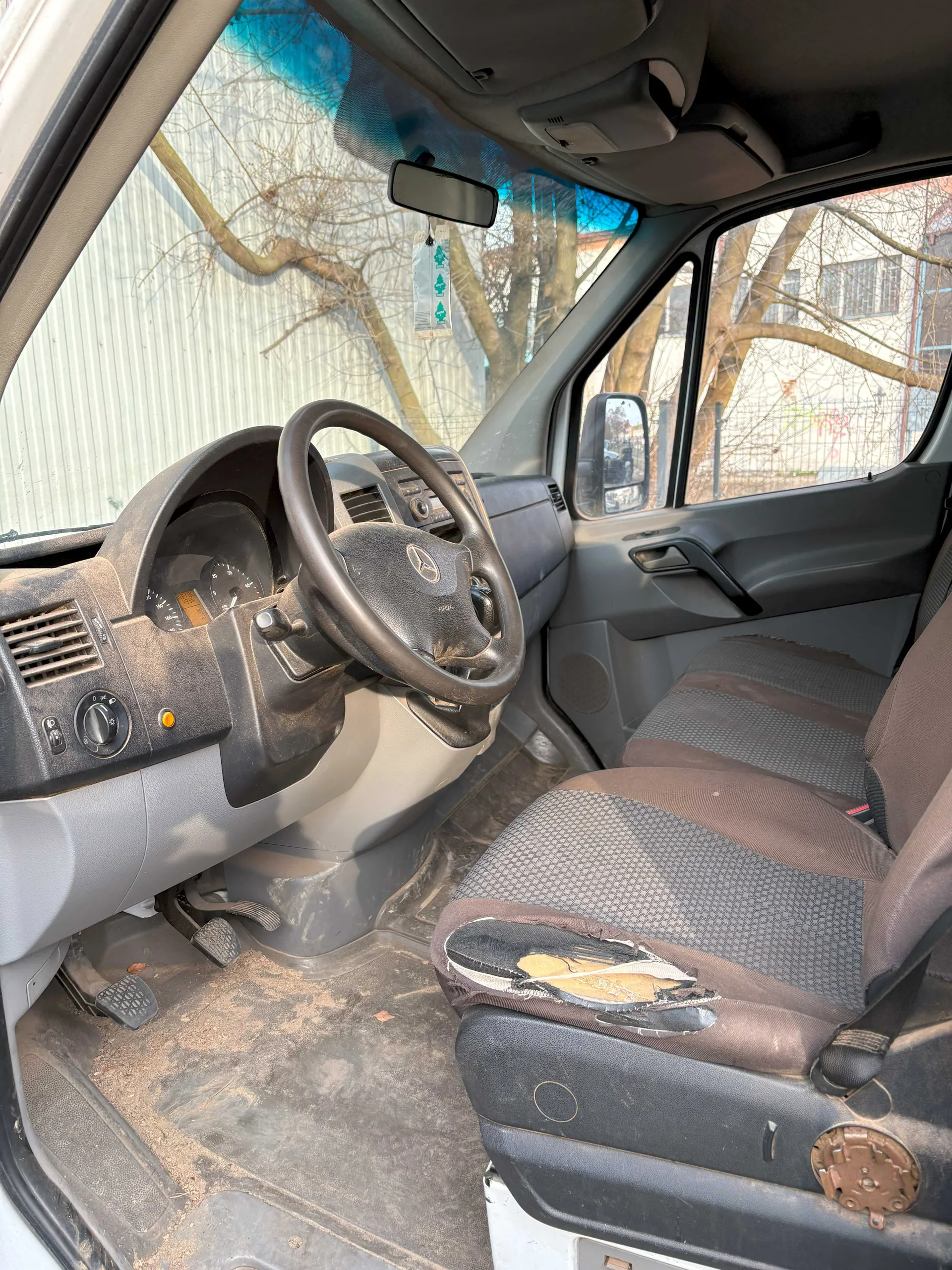 Wnętrze samochodu dostawczego marki Mercedes-Benz. Widoczna kierownica z logo, zniszczona siedzisko kierowcy z odsłoniętym podkładem, brudna podłoga z kurzem i śmieciem, deska rozdzielcza z licznikami i przyciskami, lusterko wsteczne z wiszącym zielonym odświeżaczem powietrza w kształcie drzewek. Przez przednią szybę widoczne drzewa bez liści i budynek z blachy falistej. Drzwi pasażerskie są otwarte, a zewnętrzne lusterko boczne odbija fragment ogrodzenia.