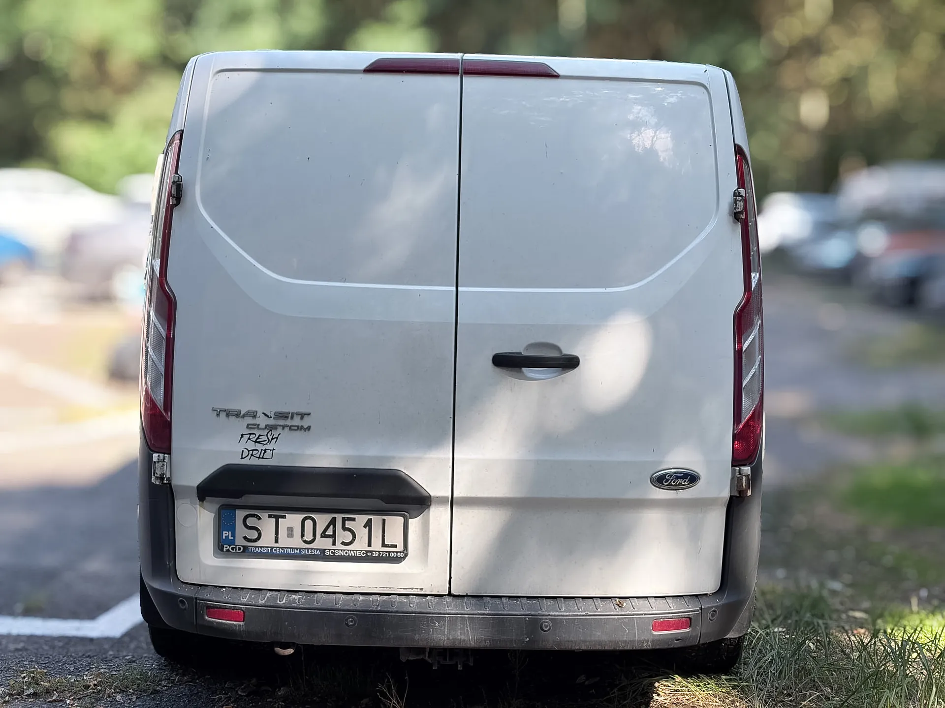Tylny widok białego vana marki Ford Transit Custom. Pojazd posiada dwie tylne klapy, czarny zderzak z czerwonymi odblaskami oraz tablicę rejestracyjną o numerze ST 0451L. Po lewej stronie widnieje napis 'TRANSIT CUSTOM' oraz odręczny napis 'FRESH DRIVE'. Po prawej stronie znajduje się logo Ford.
