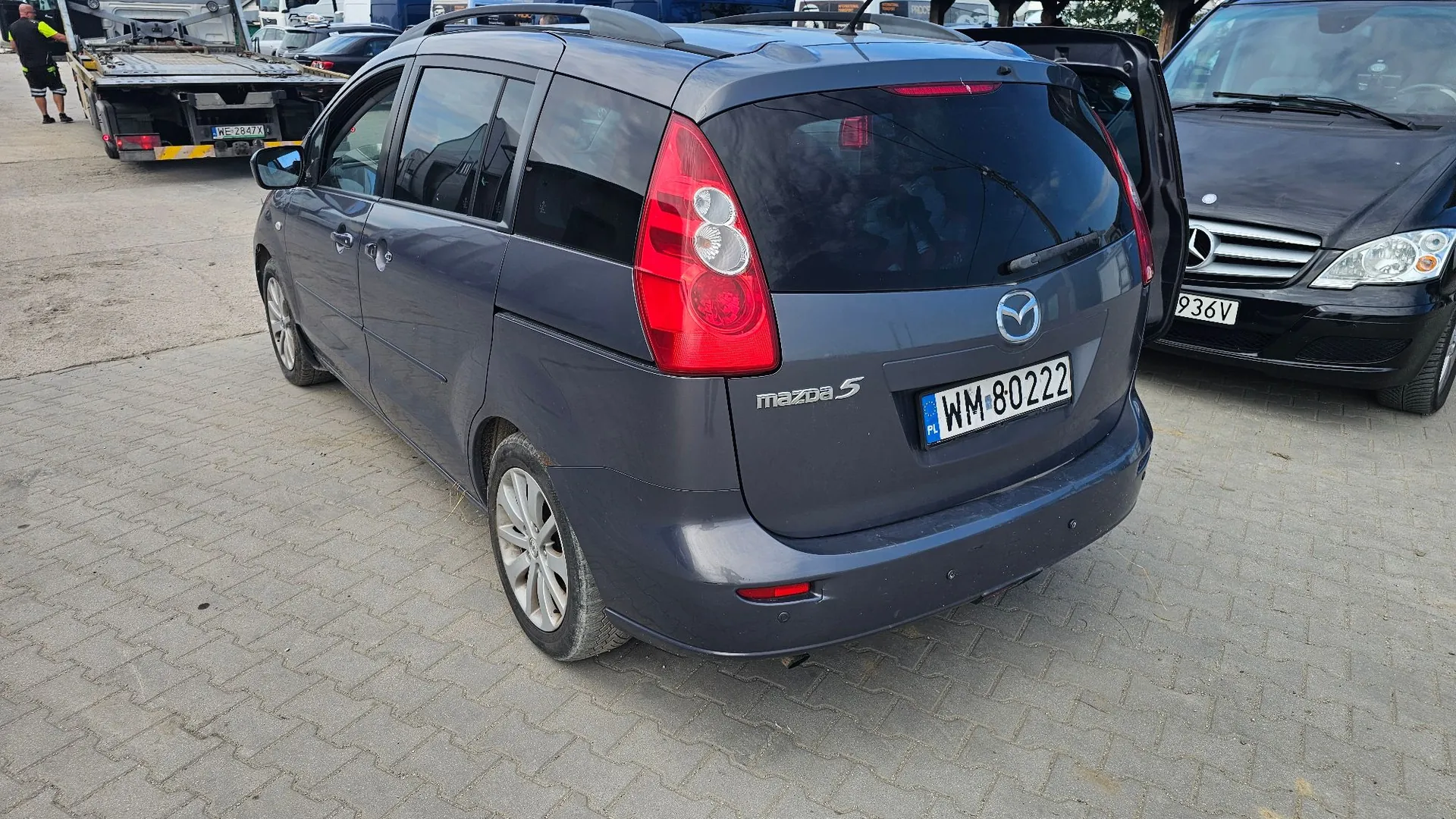 Szary samochód marki Mazda 5 zaparkowany na kostce brukowej. Widok z tyłu i z boku. Samochód ma polską tablicę rejestracyjną o numerze WM 80222. W tle widoczny jest inny ciemny samochód oraz laweta z pojazdem.