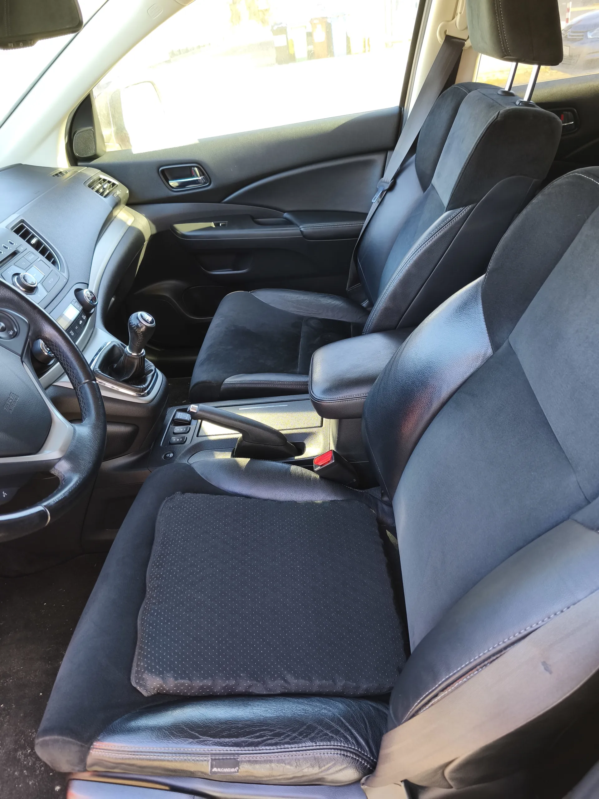 Wnętrze samochodu z czarnymi skórzano-tekstylnymi fotelami, kierownicą po lewej stronie, dźwignią zmiany biegów, centralną konsolą z przyciskami oraz drzwiami z uchwytem i sterowaniem okien. Na fotelu kierowcy znajduje się czarna poduszka z drobnym wzorem. Światło słoneczne wpada przez przednią szybę.