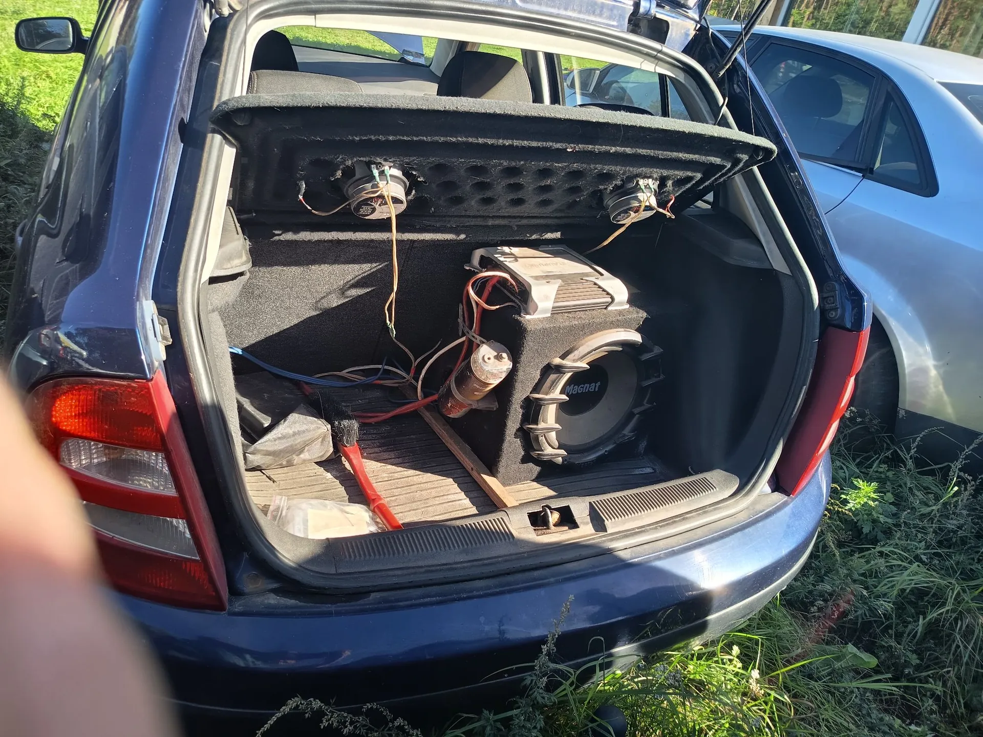 Otwarty bagażnik ciemnoniebieskiego samochodu, w którym zamontowano system audio. Wewnątrz znajduje się czarna obudowa z głośnikiem niskotonowym marki Macnat, na której wierzchu spoczywa wzmacniacz z widocznymi przewodami. W podniesionej klapie bagażnika zamontowane są dwa mniejsze głośniki. W tle widoczny jest fragment srebrnego samochodu oraz trawa.
