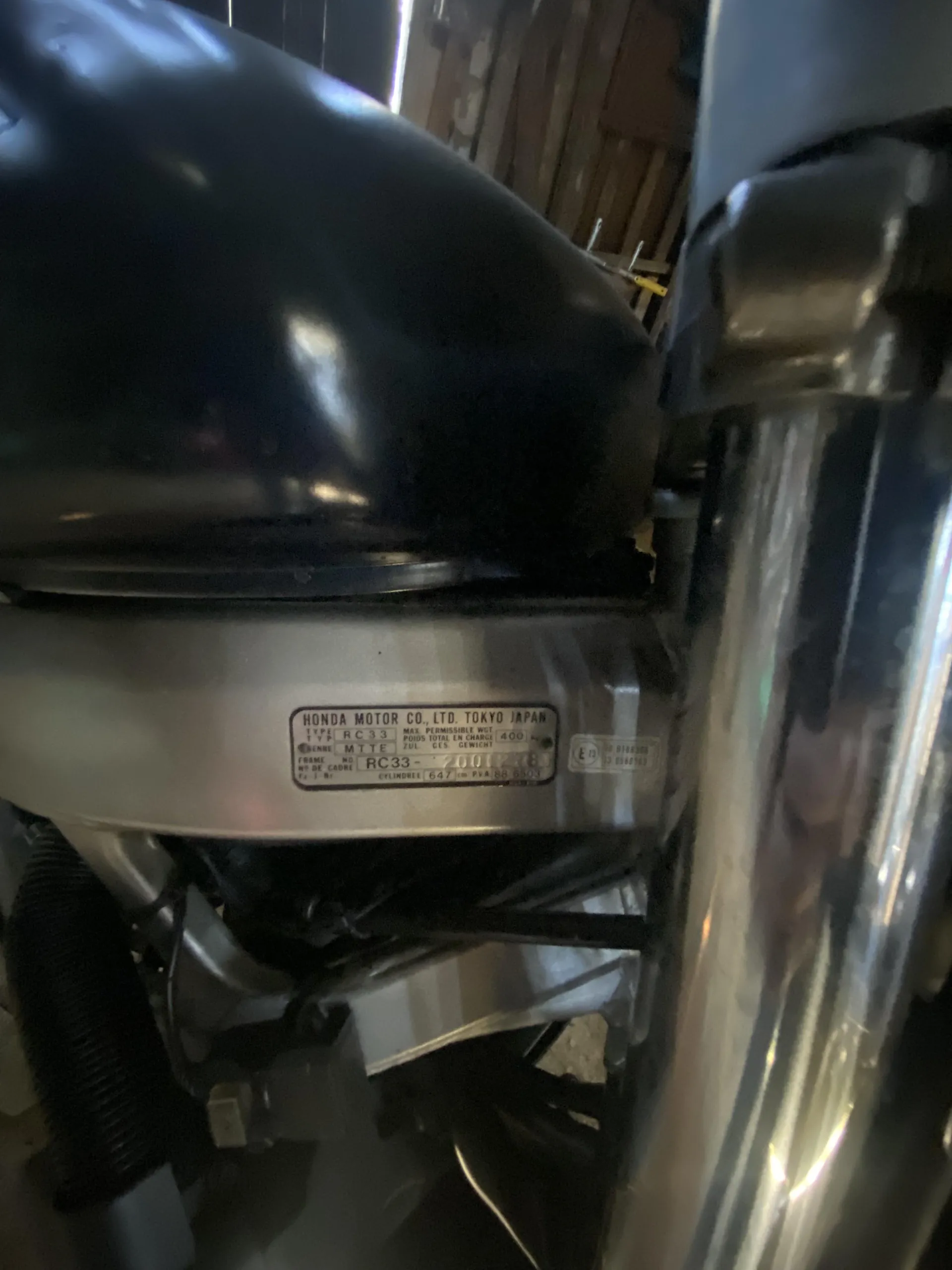 Zbliżenie na metalową tabliczkę z napisem 'HONDA MOTOR CO., LTD. TOKYO JAPAN', poniżej widoczne dane techniczne: 'TYPE RC33', 'MAX. PERMISSIBLE WT. 400', 'POIDS TOTAL EN CHARGE', 'ZUL. GES. GEWICHT', 'FRAME NO. RC33-2001278', 'CYLINDRE 647 cm³ P/N 88 6503'. Tabliczka zamontowana na metalowej konstrukcji motocykla, w tle częściowo widoczne elementy garażu i narzędzia.