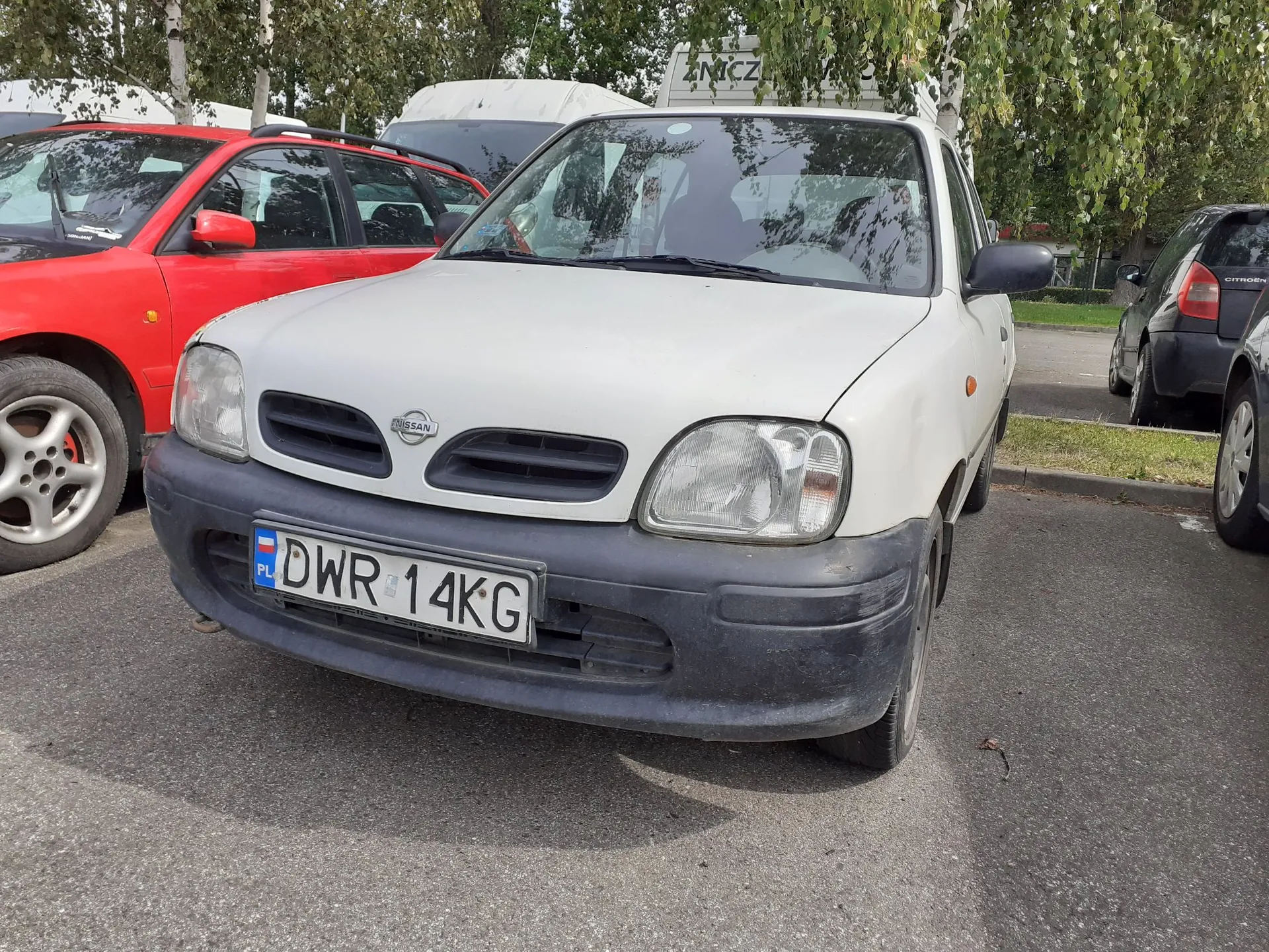 Biały samochód marki Nissan zaparkowany na asfaltowym placu. Pojazd ma czarny plastikowy zderzak z polską tablicą rejestracyjną o numerze DWR 14KG. Po lewej stronie widoczny jest fragment czerwonego samochodu, a po prawej ciemny samochód. W tle znajdują się zielone drzewa i biały pojazd dostawczy.